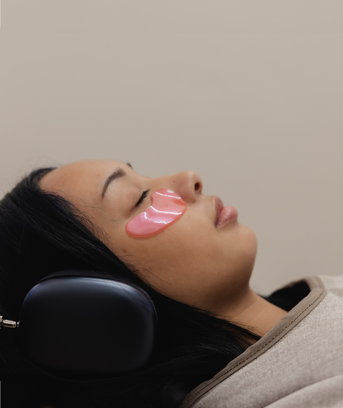 Woman with closed eyes lying down wearing pink under-eye gel patches and black headphones.