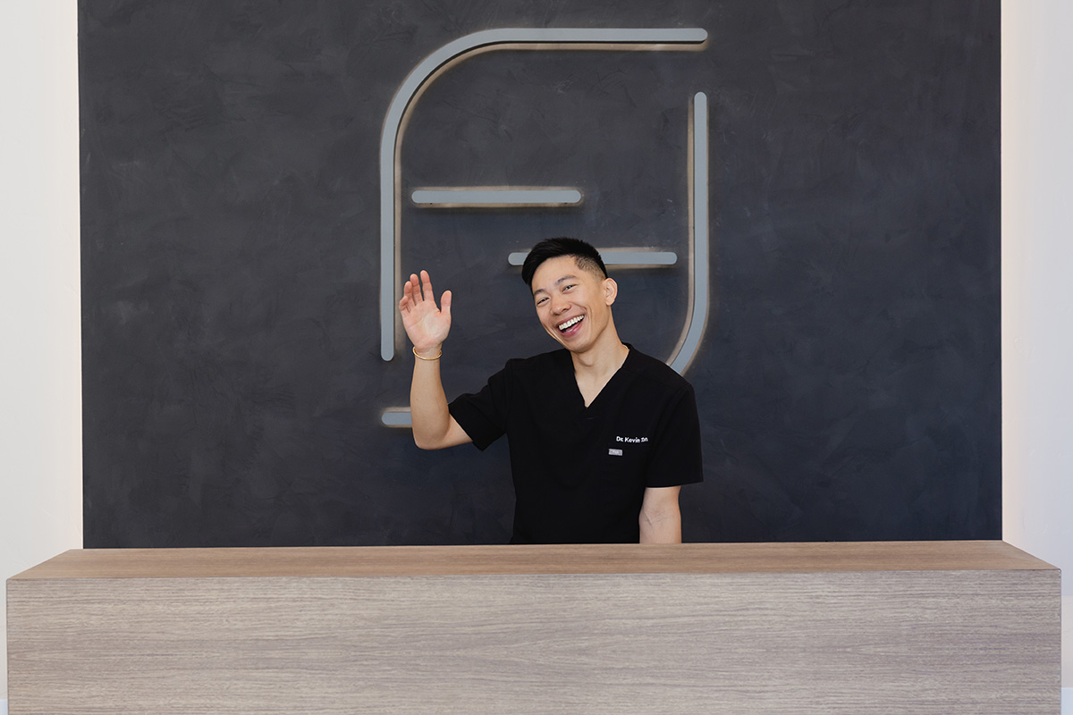 Smiling man in black scrubs waving behind a wooden reception desk with a modern wall logo.