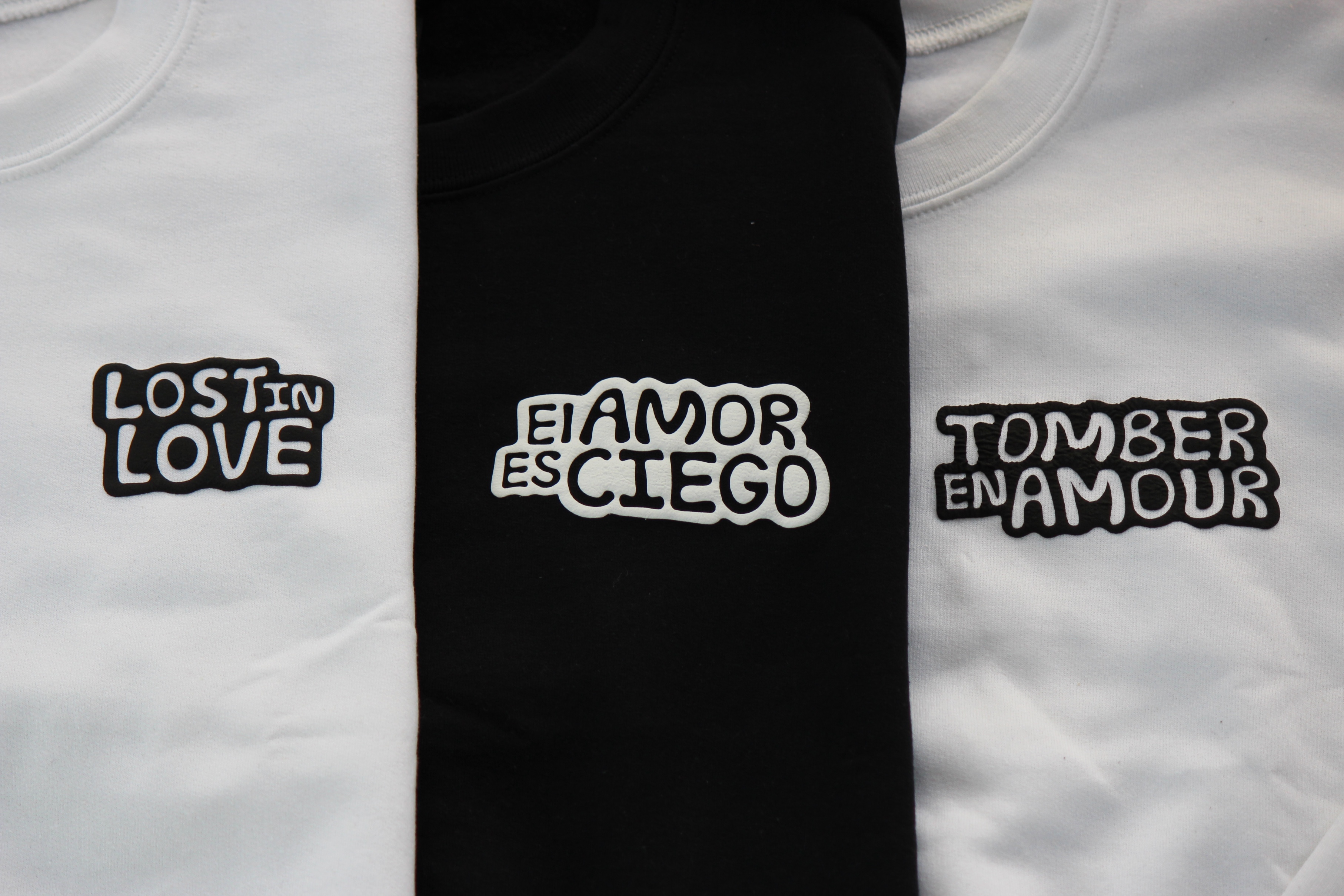 Three folded t-shirts with love phrases: Lost in Love, El Amor es Ciego, and Tomber en Amour.