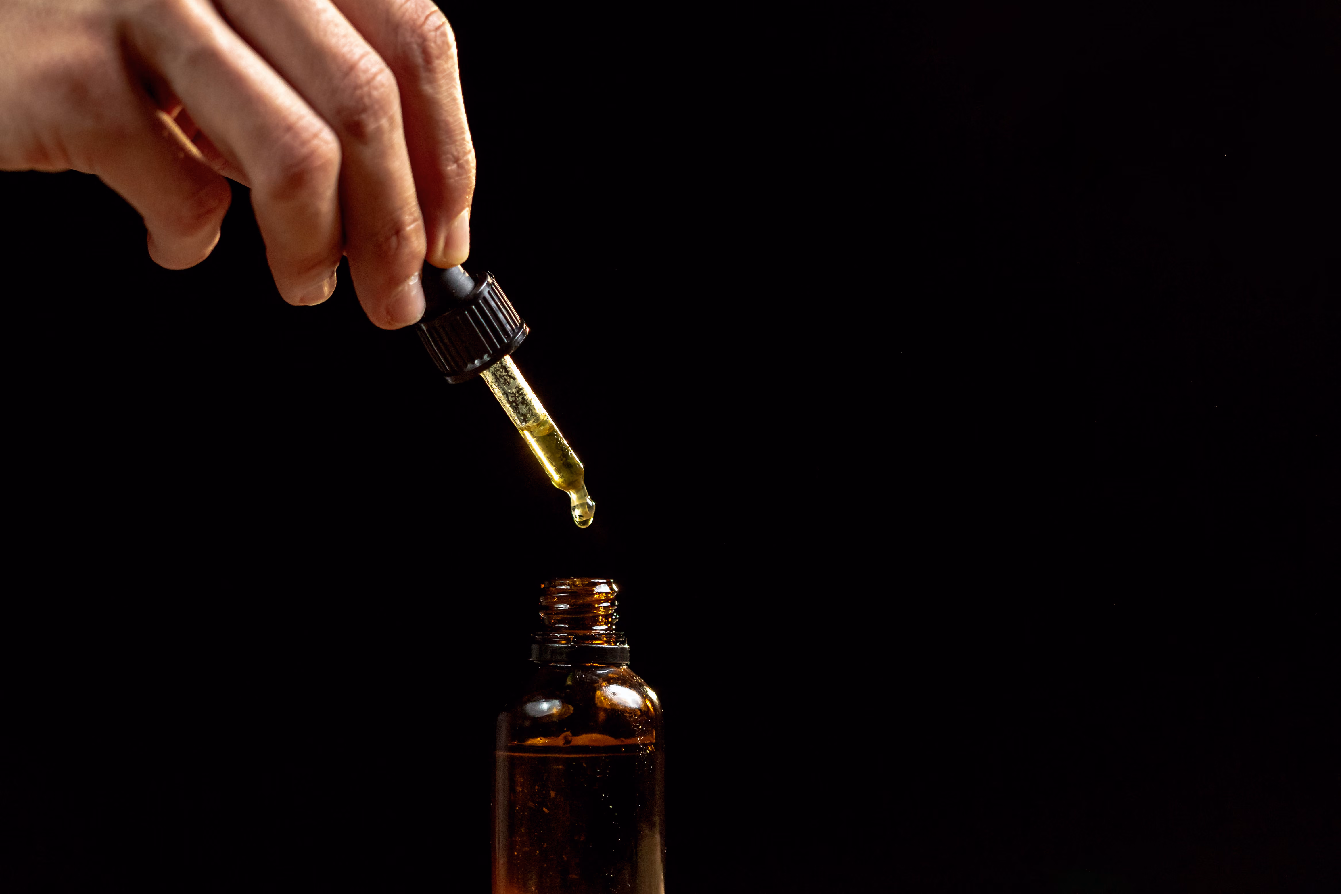 Hand holding a dropper releasing a drop of yellowish liquid into a small brown glass bottle against a black background.