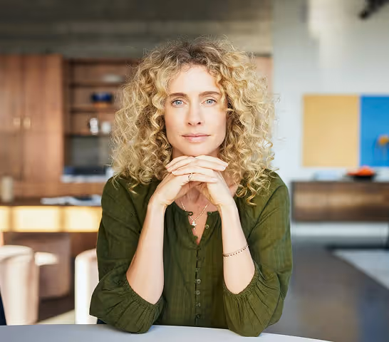 Woman with curly blonde hair wearing a green shirt, sitting indoors with her hands clasped under her chin, looking directly at the camera.