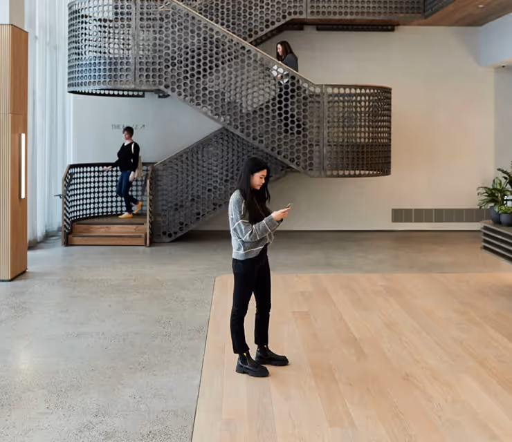 Three people in a modern interior with perforated metal staircase; one woman standing on wooden floor looking at her phone.