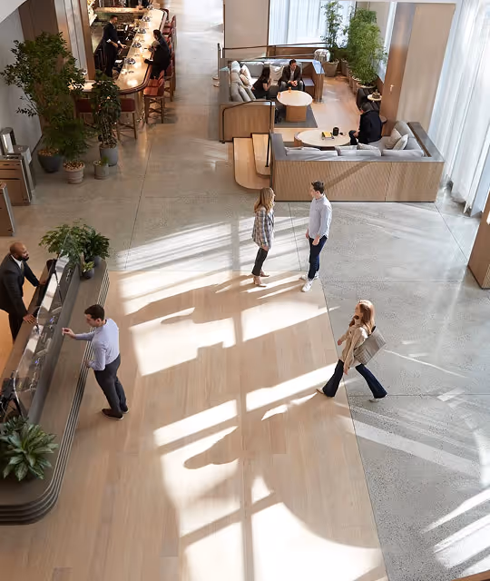 Modern office lobby with people interacting near reception and sitting in lounge areas with large windows casting shadows on the floor.