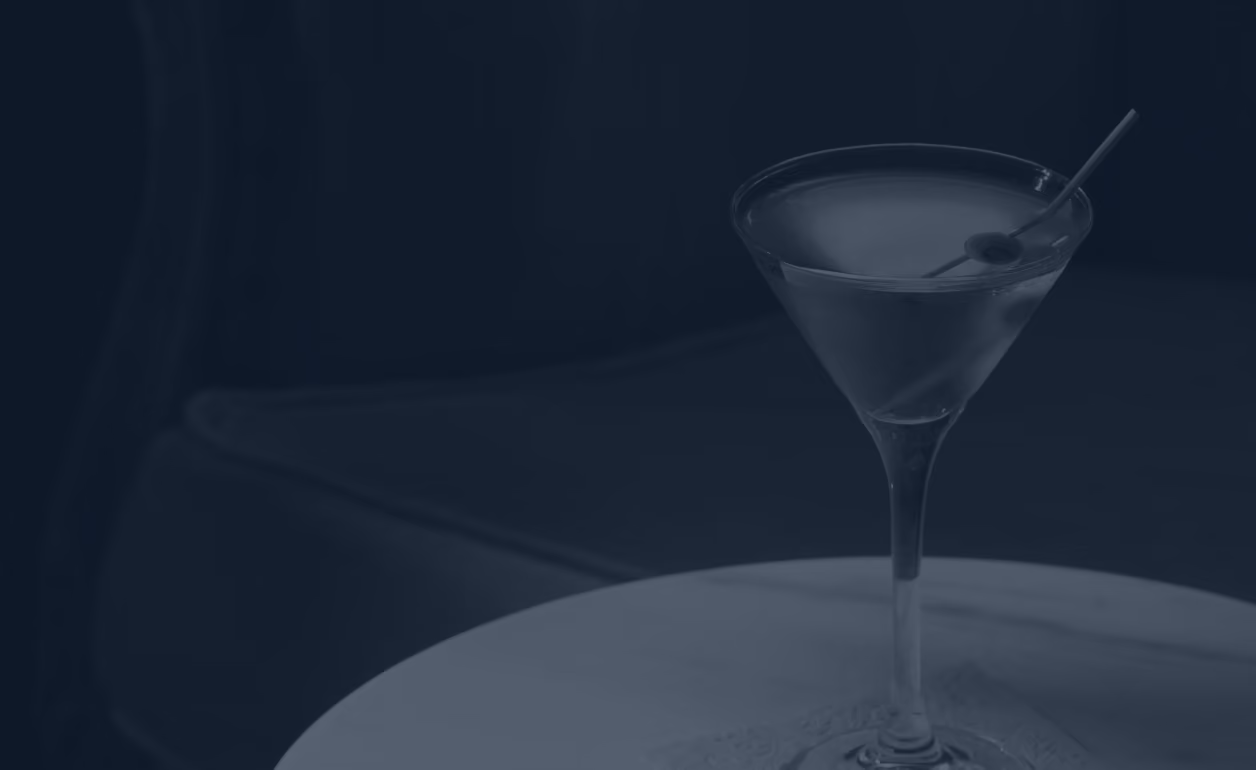 A martini glass with an olive garnish on a table against a dark blue background.
