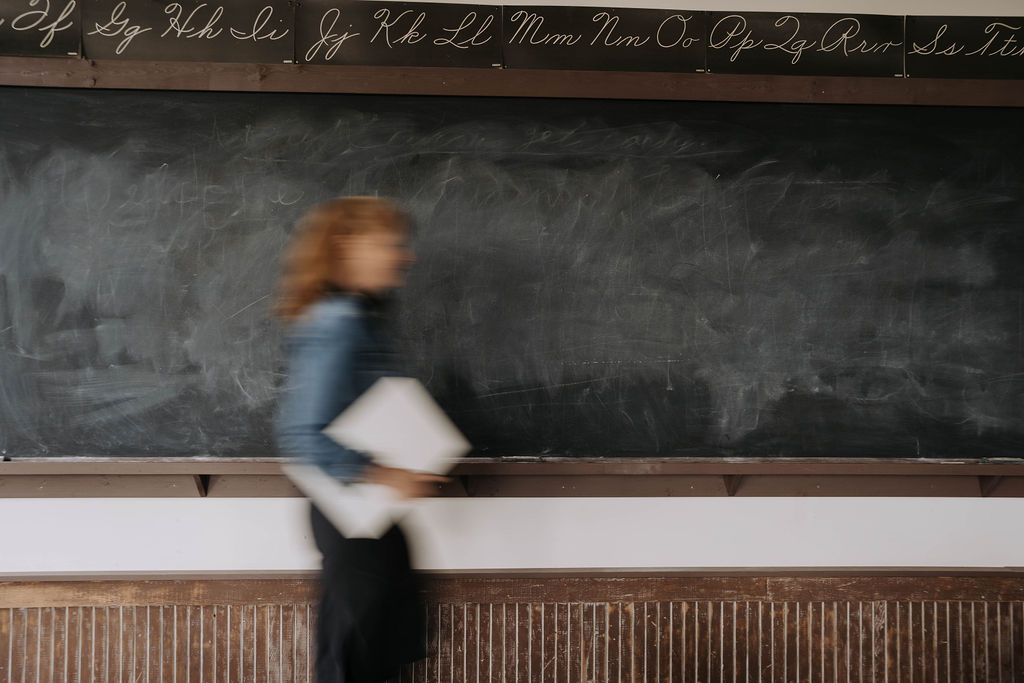 Kim Thompson in motion, carrying a laptop and papers past a chalkboard in a vintage schoolhouse classroom.