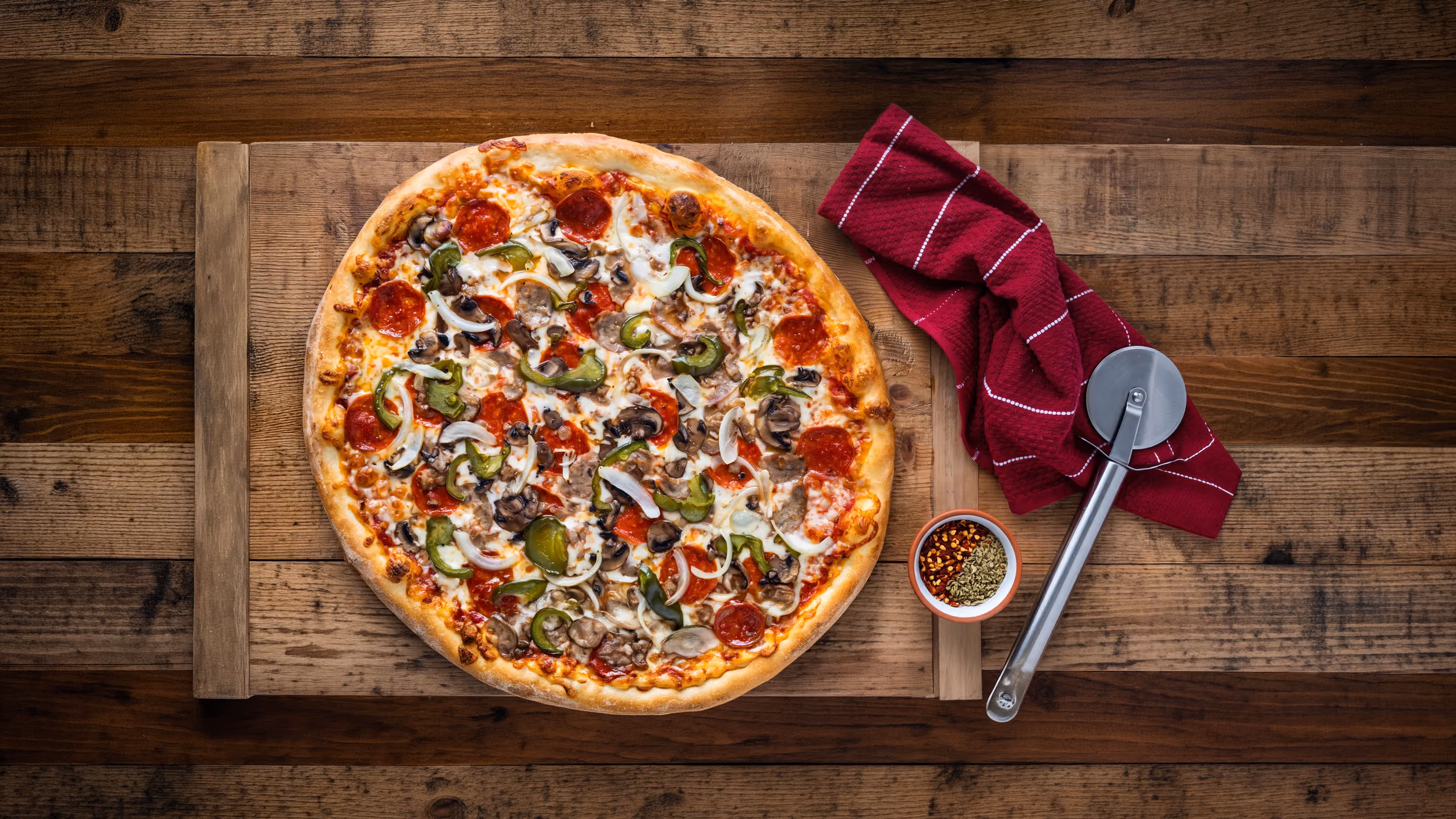 Pepperoni pizza with mushrooms, onions, and green peppers on a wooden board with a pizza cutter and red towel.