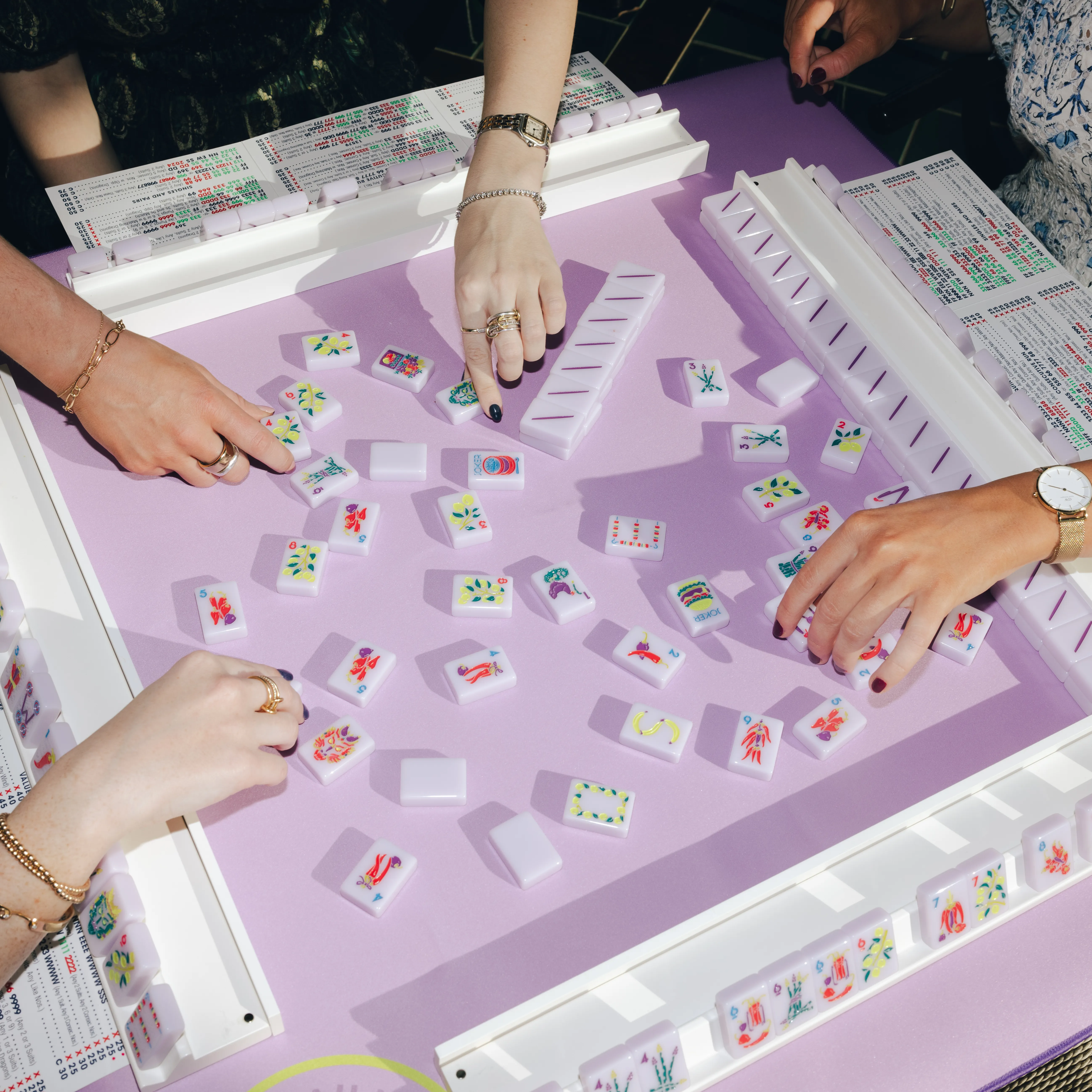Hands playing mahjong on a lavender table with colorful tiles