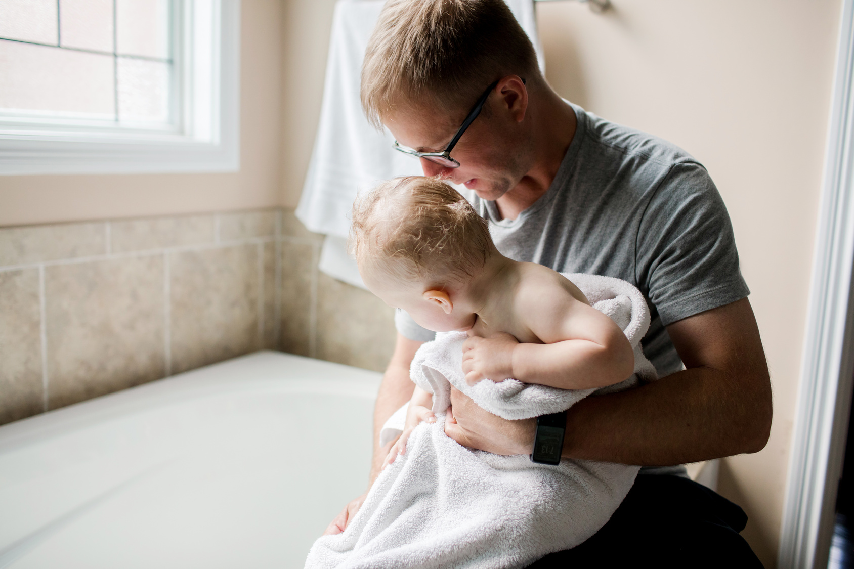 Father holding and drying a baby wrapped in a towel near a bathtub in a bathroom.