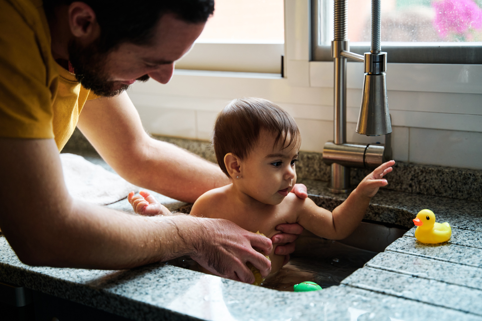 Father bathing toddler in kitchen sink while child reaches for a yellow rubber duck.