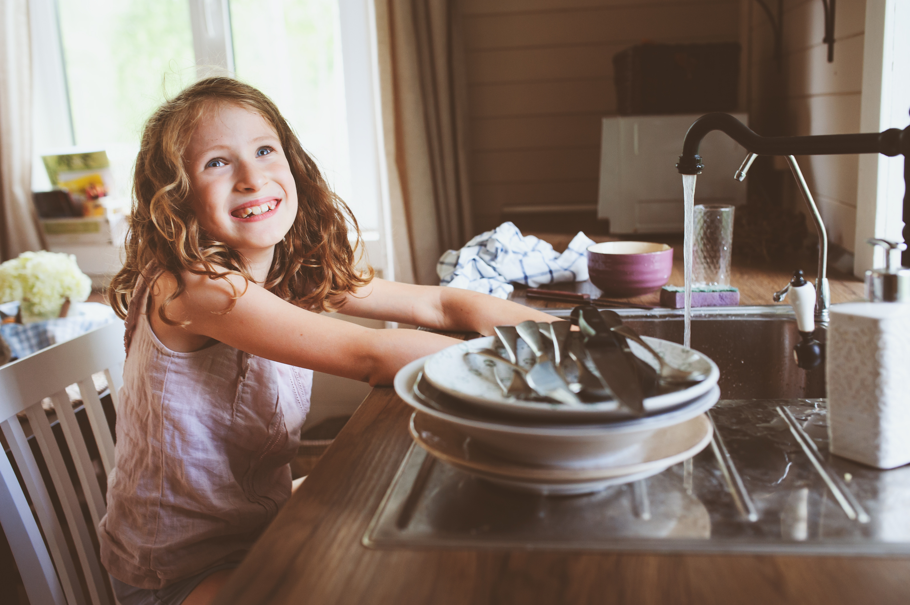 Smiling girl with curly hair washing dishes at kitchen sink with water running from faucet.