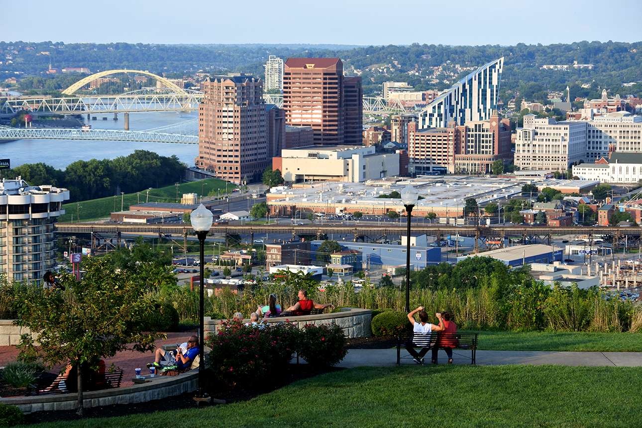 People sitting on benches on a hill overlooking a city skyline with bridges and a river.