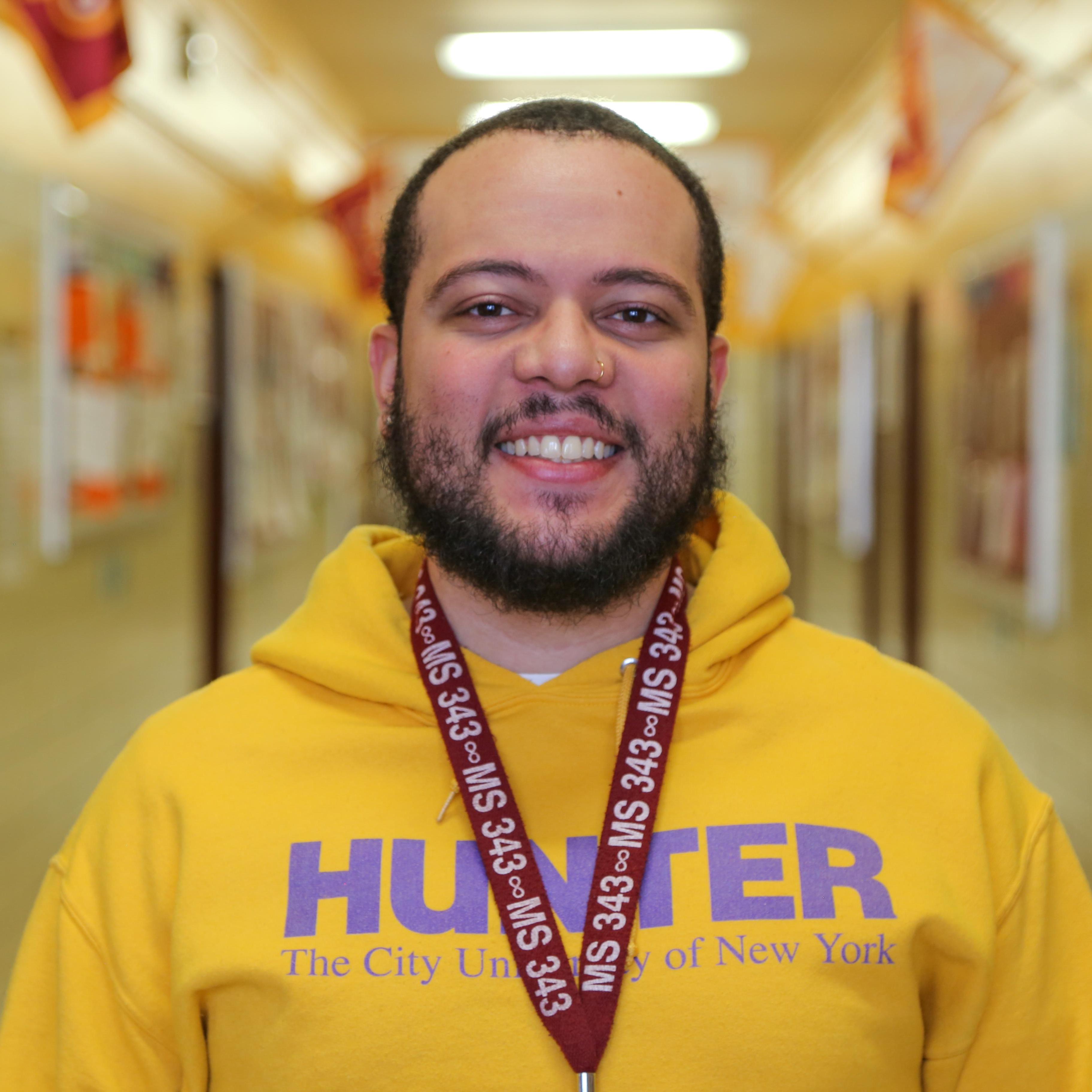 Mr Batista smiling in school hallway wearing Hunter College sweatshirt