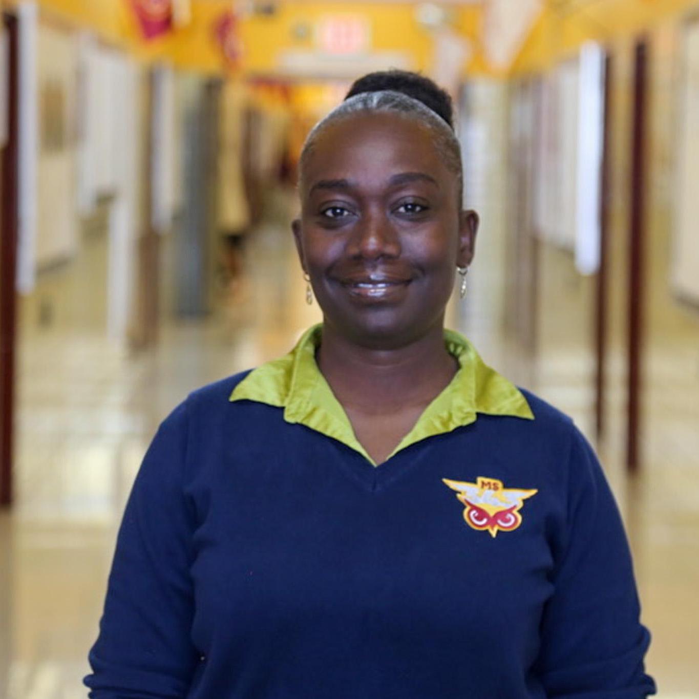 Ms. Cooper smiling in school hallway wearing staff uniform