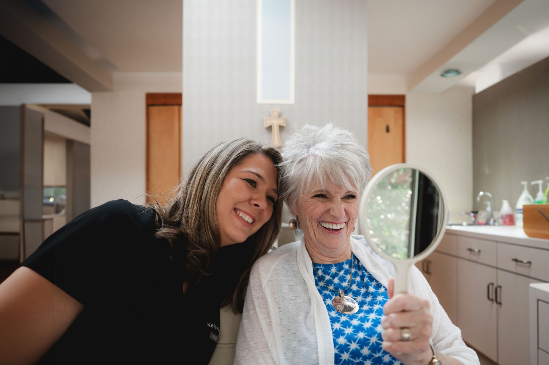 Dental team member sharing a laugh with a smiling senior patient holding a hand mirror.