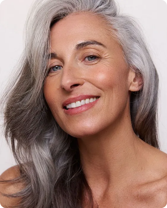 Close-up of a smiling woman with long gray hair and clear skin against a plain background.