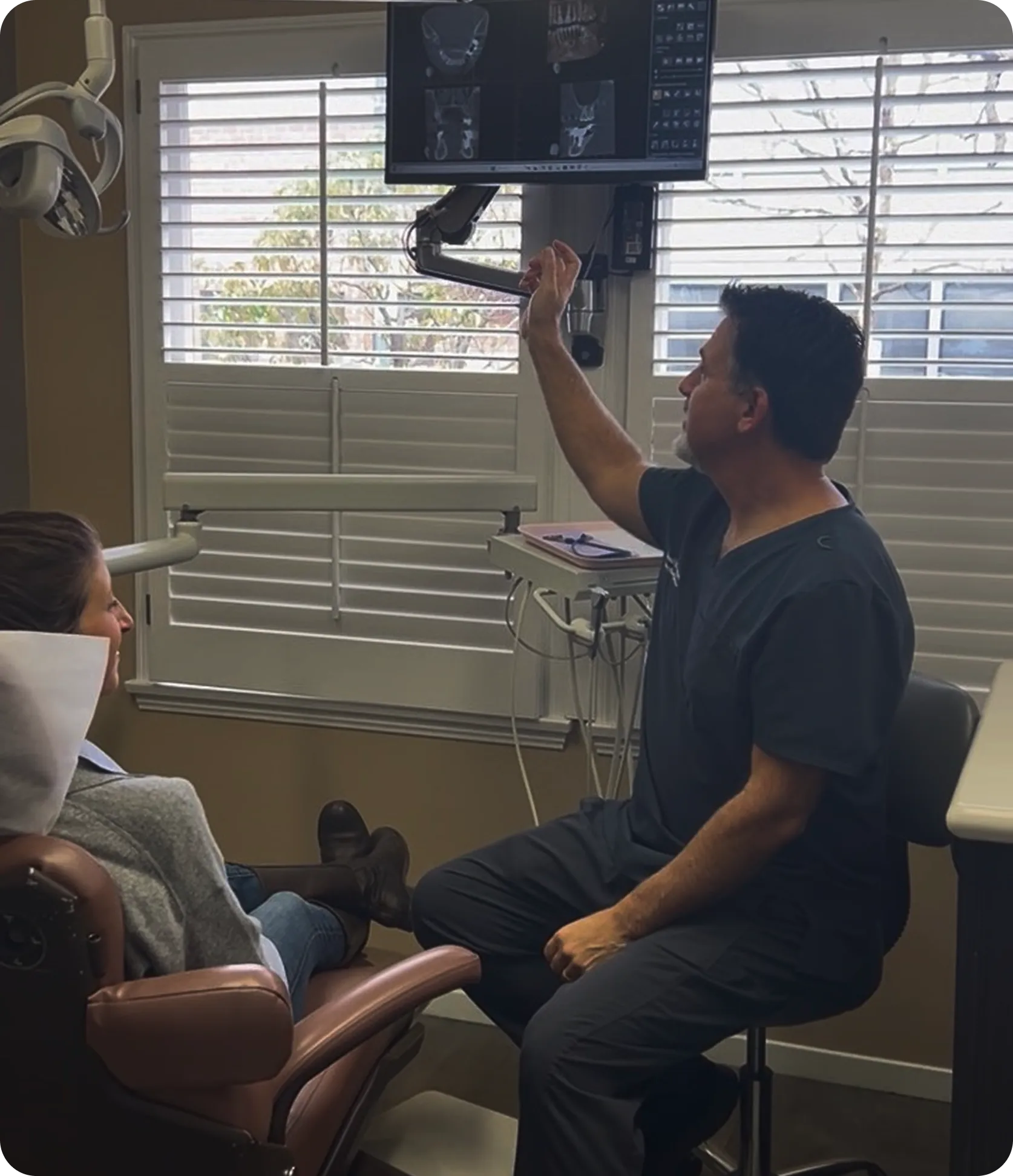 Dentist showing dental X-rays on screen to patient seated in chair in office with window blinds.