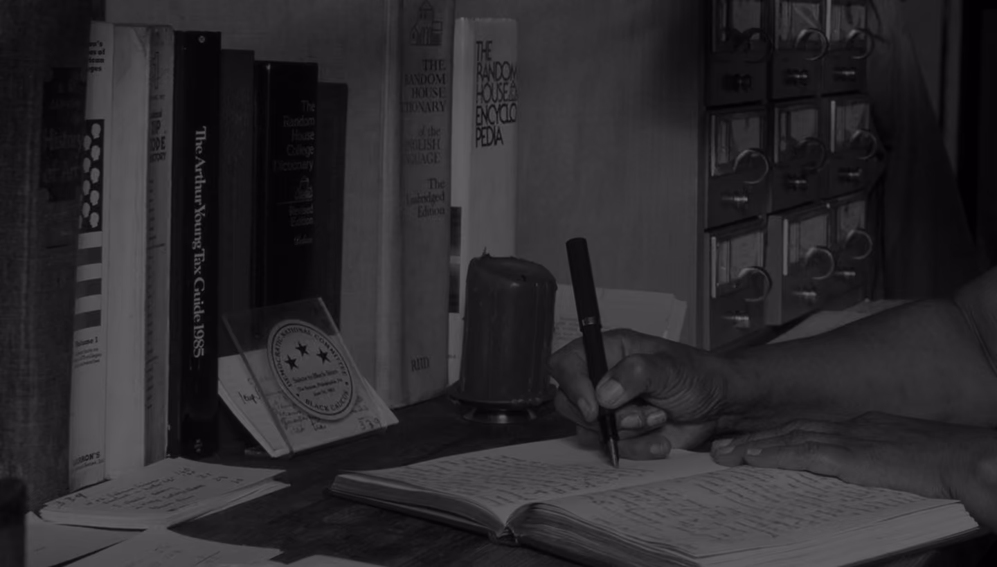 A person writing notes with a pen in an open notebook on a wooden desk, with a shelf of books and a card catalog in the background.