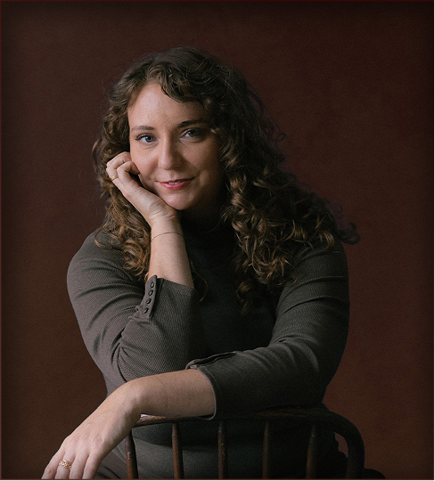 A woman with curly brown hair and light skin sitting on a wooden chair facing backward, resting her head on her right hand, wearing a long-sleeve dark green top, with a calm expression against a dark brown background.