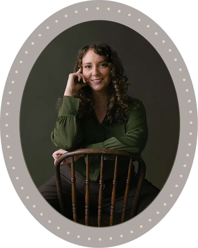 A woman with curly brown hair wearing a green blouse sits on a wooden chair turned backward against a plain dark background, smiling with her head resting on her hand.
