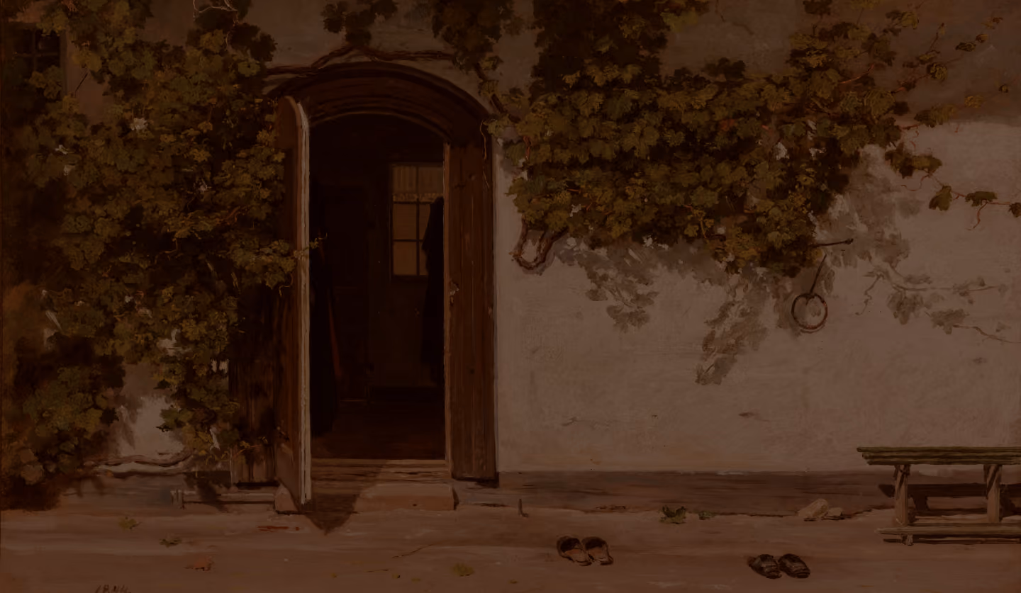 Open wooden door in a white wall covered with leafy vines, showing a dark interior with a window, and pairs of shoes on the ground outside.