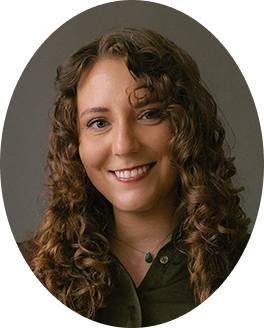 Portrait of a young woman with curly brown hair, smiling and wearing a dark green top and a necklace with a dark pendant against a gray background.