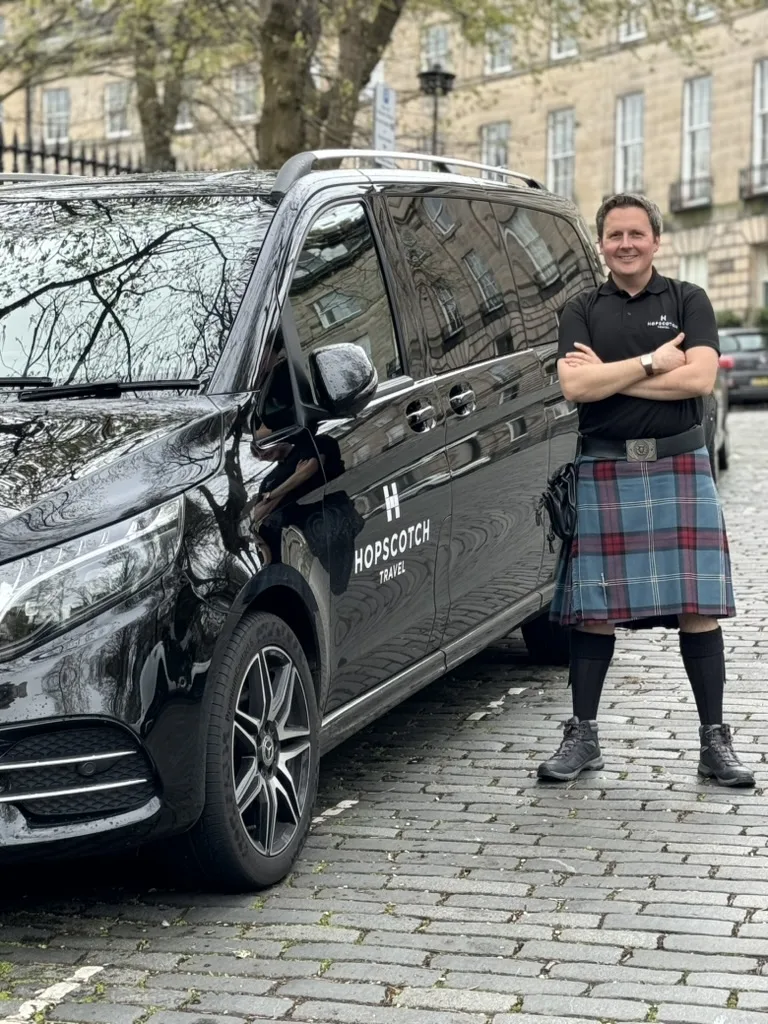 Man in kilt standing beside black Hopscotch Travel tour van in Scottish city square.