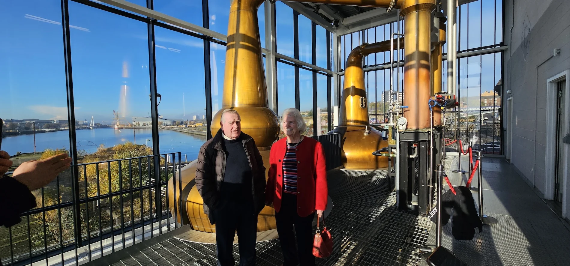 Visitors inside Scottish whisky distillery with golden copper stills and river views.