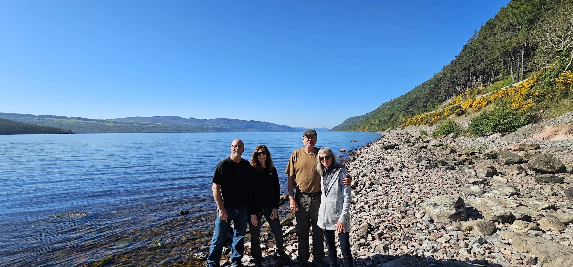 Four tourists posing on a rocky beach beside a Scottish loch with forested hills