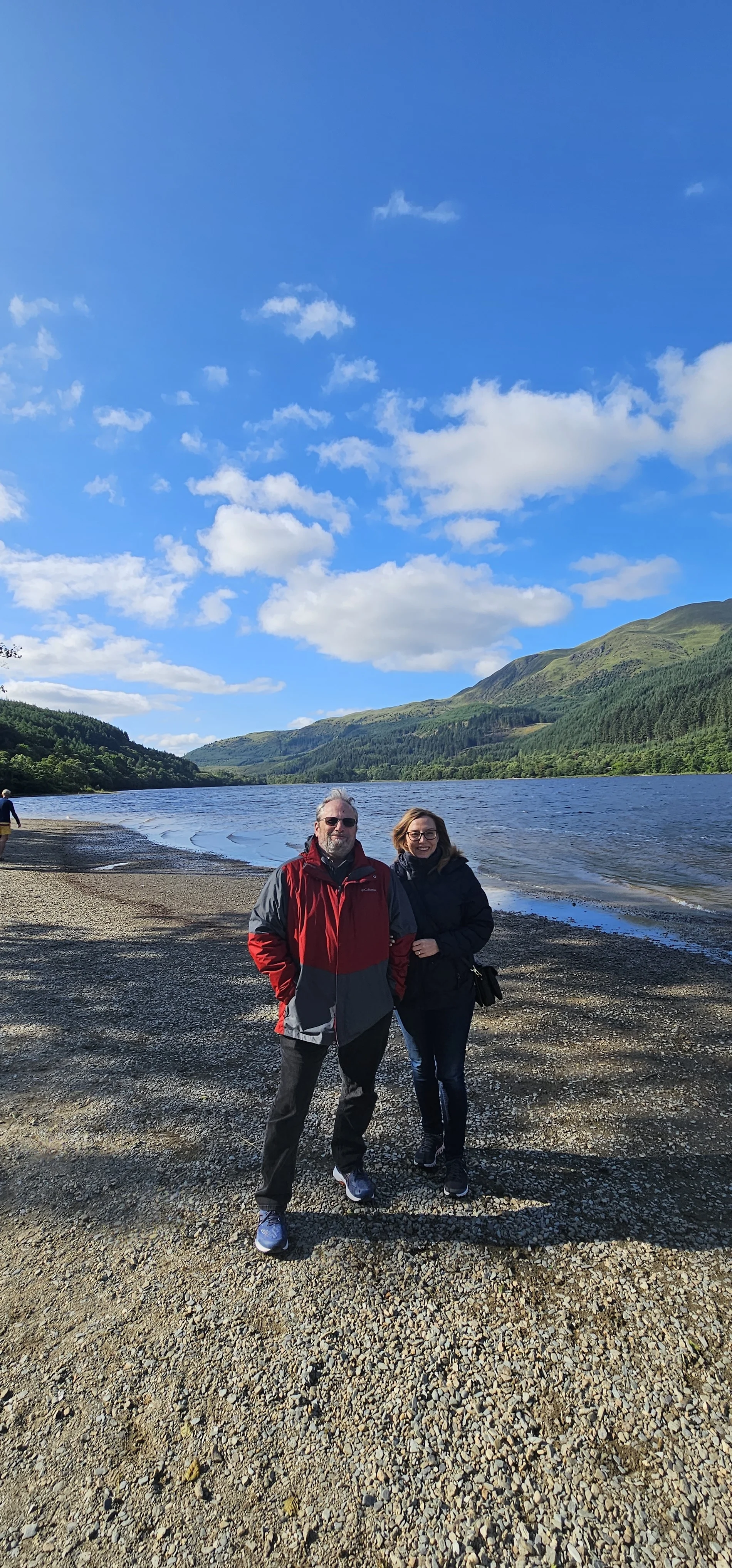 Couple posing on Scottish loch beach with forested mountains under blue sky