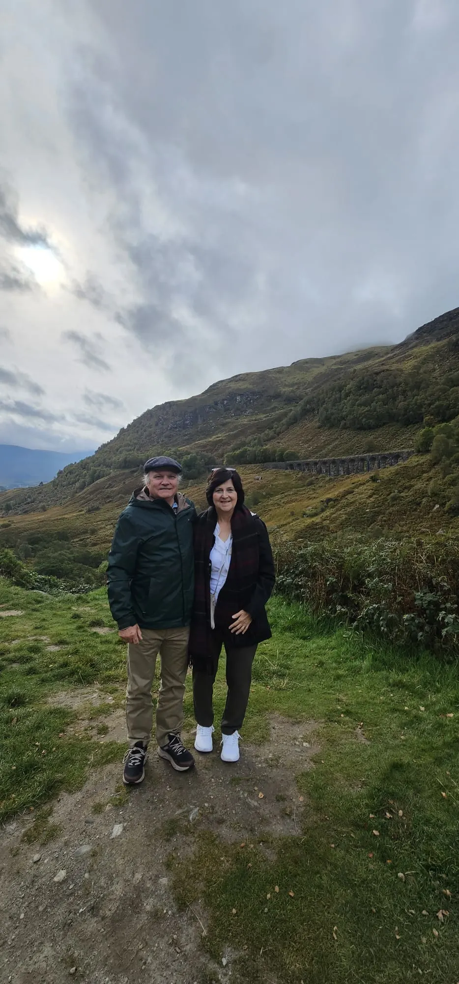 Two tourists posing in Scottish Highlands valley with mountains and cloudy sky.