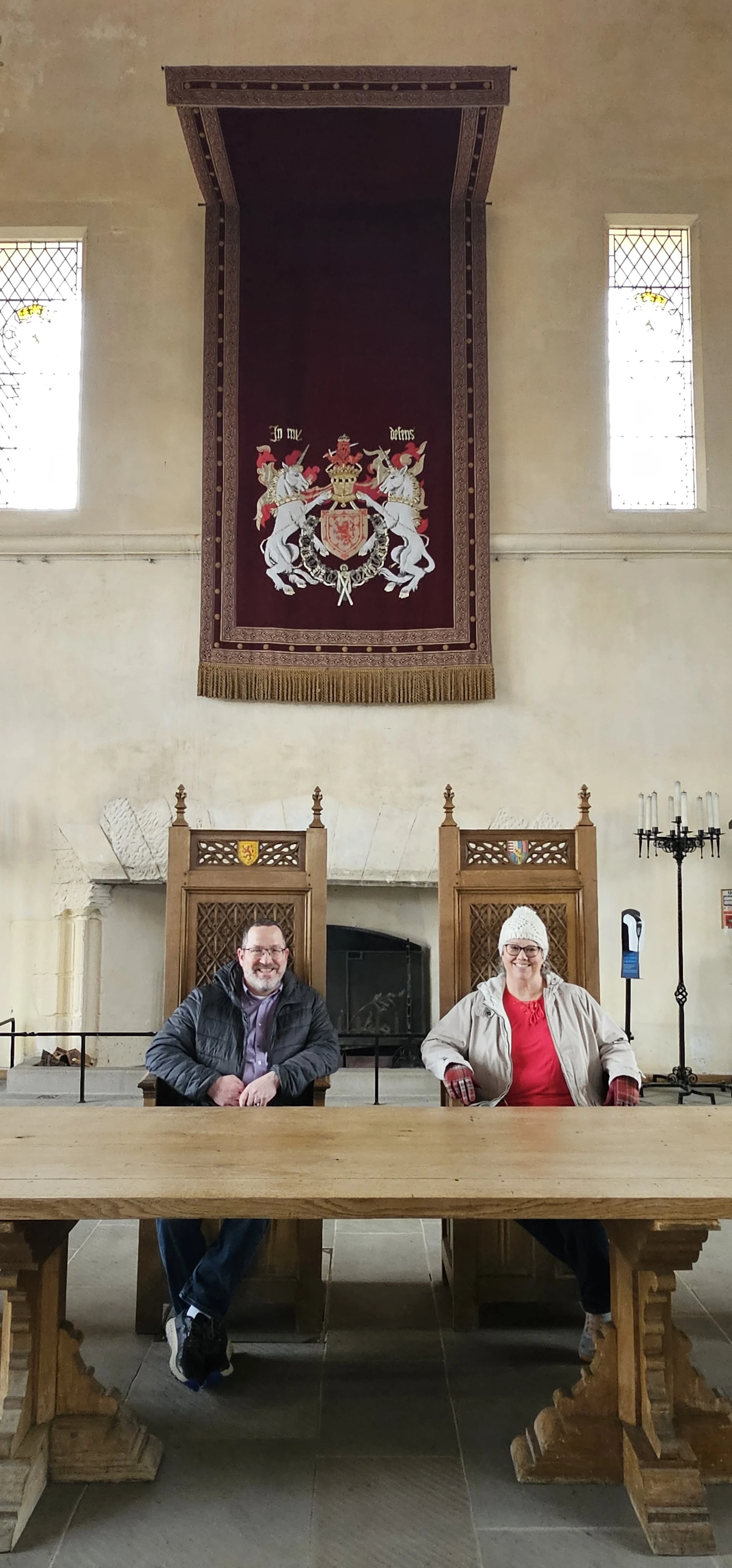 Two people sit at a wooden table inside a Scottish castle hall with heraldic banner.