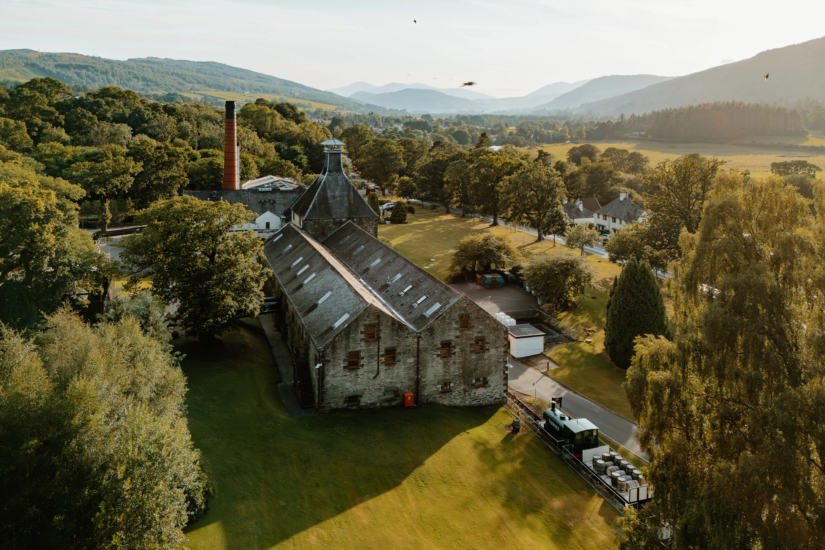 Historic Scottish whisky distillery in Highland valley with mountains and river.