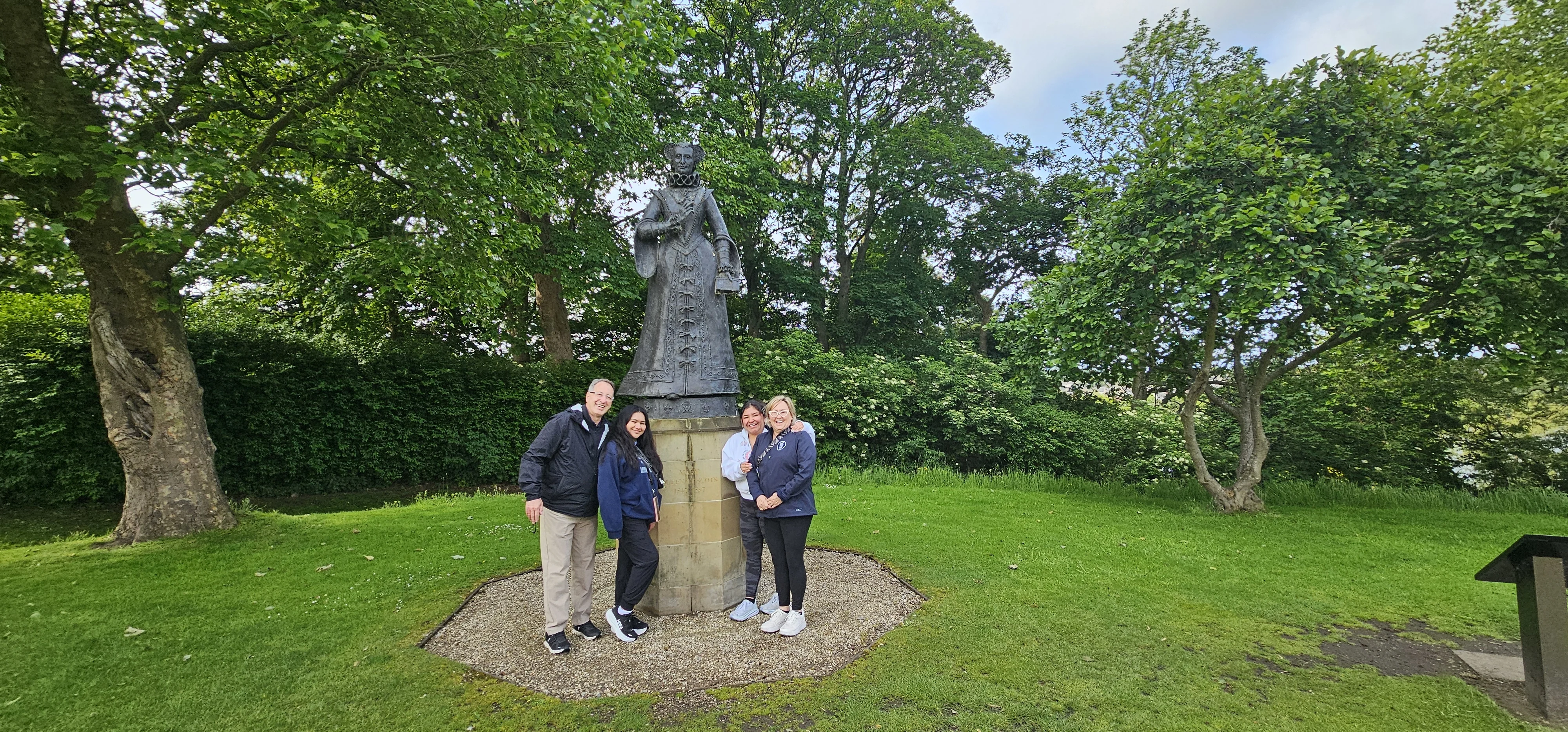 Four tourists posing beside a stone statue in a lush Scottish garden.