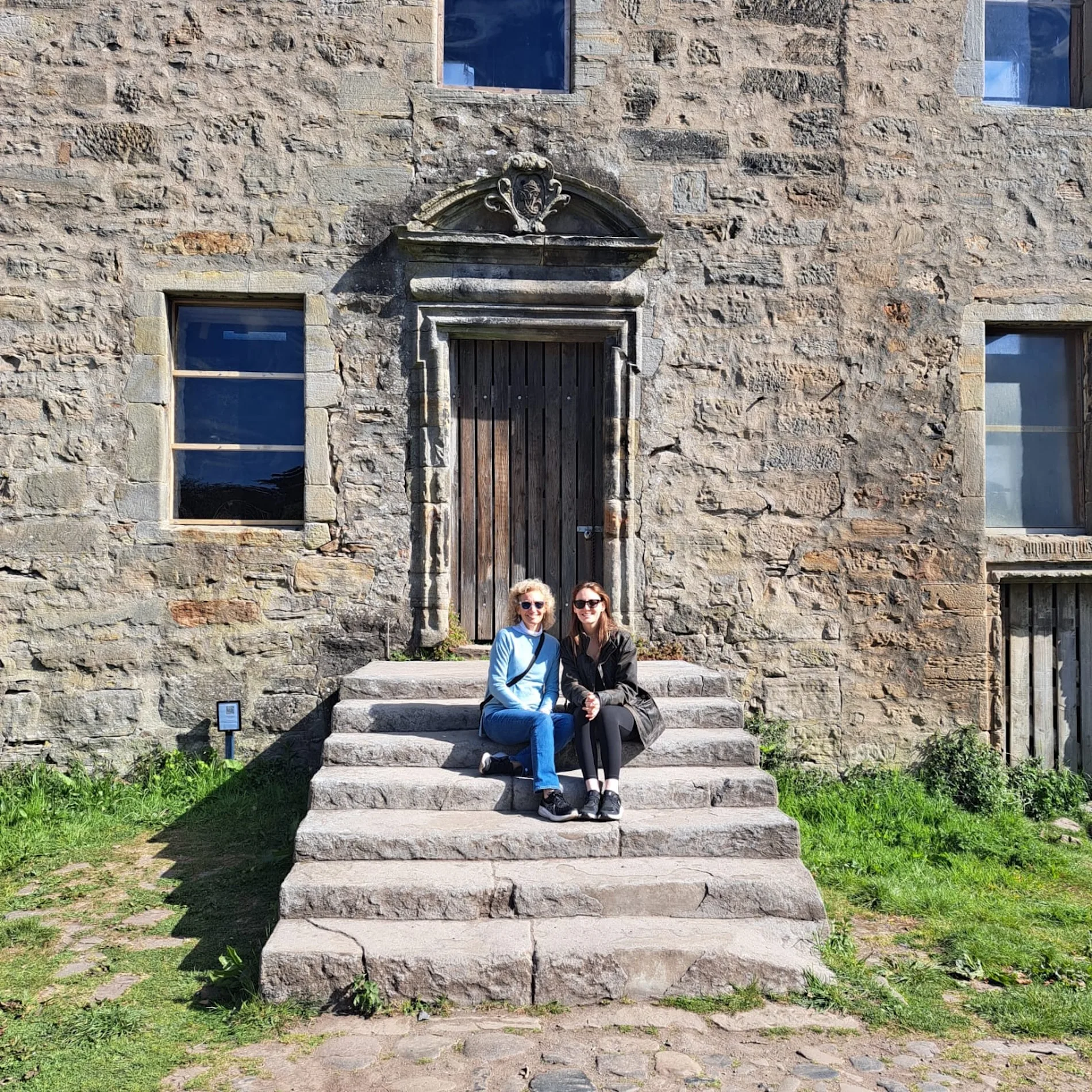 Two tourists sitting on steps of a historic Scottish stone building entrance.