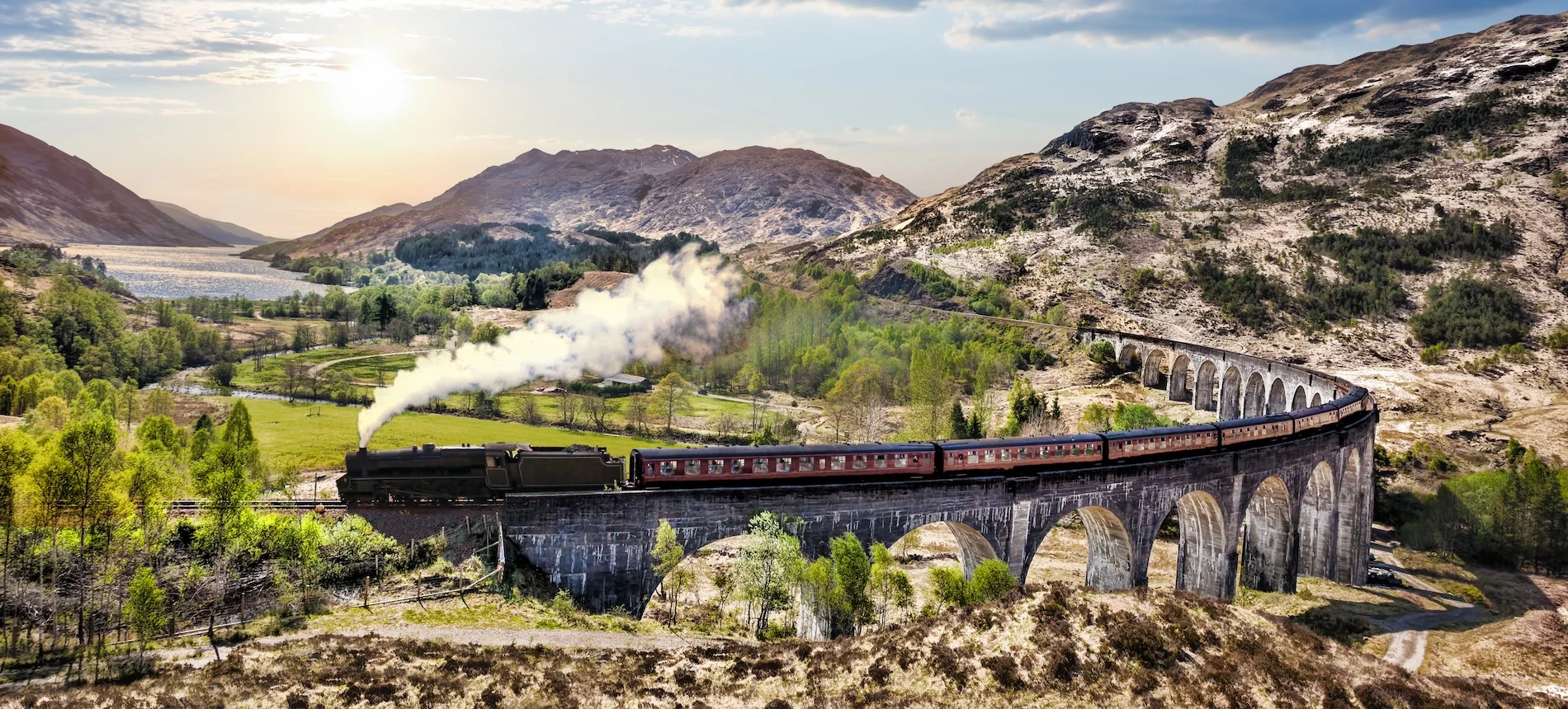 Steam train crossing stone viaduct through Scottish Highland mountains and valley.