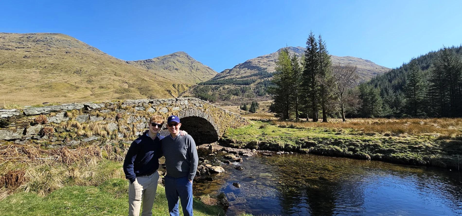 Two visitors posing by a stone bridge over a Highland stream with mountains and forest
