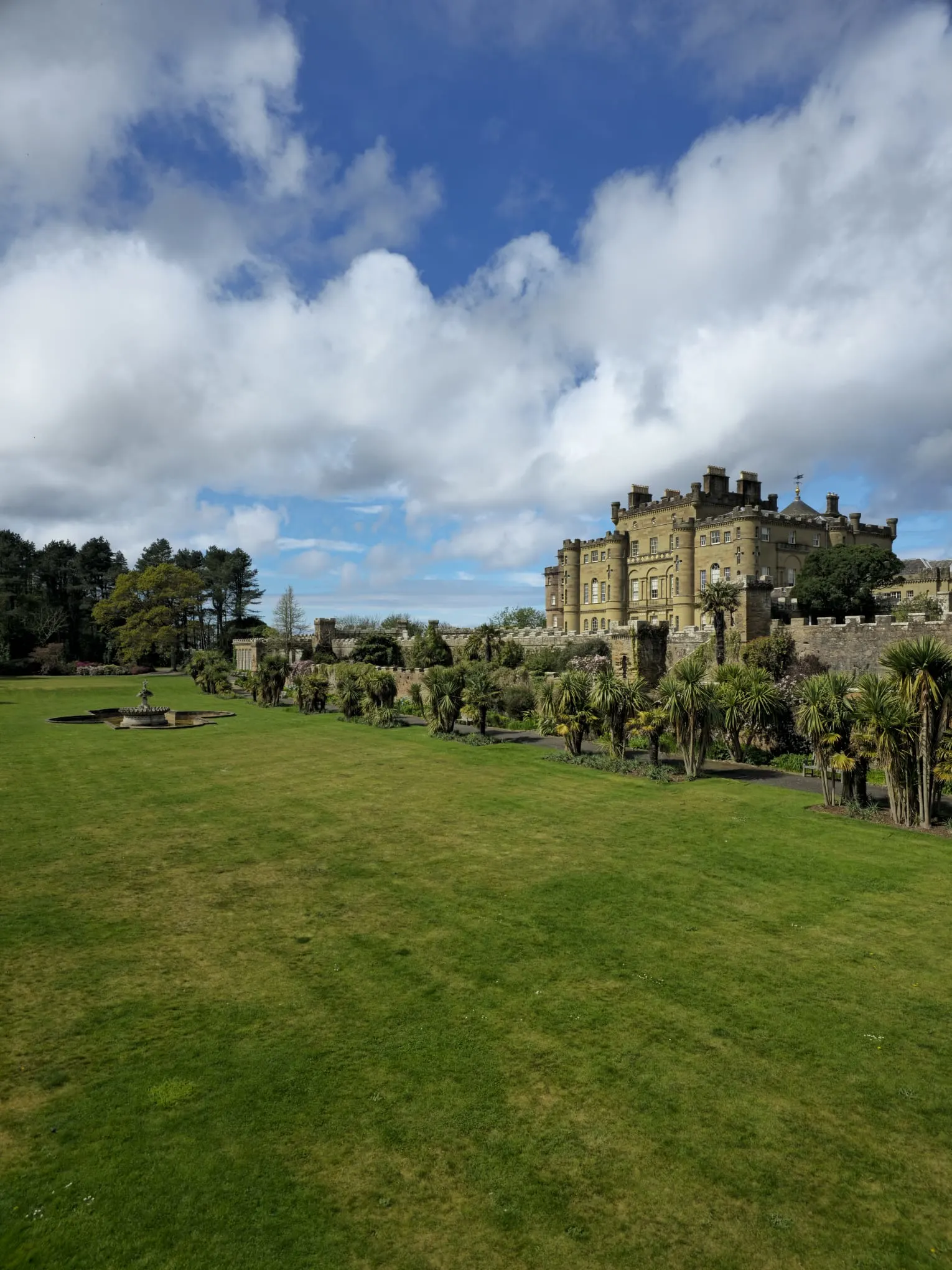 Scottish castle with manicured grounds, palm trees, and blue sky with clouds.