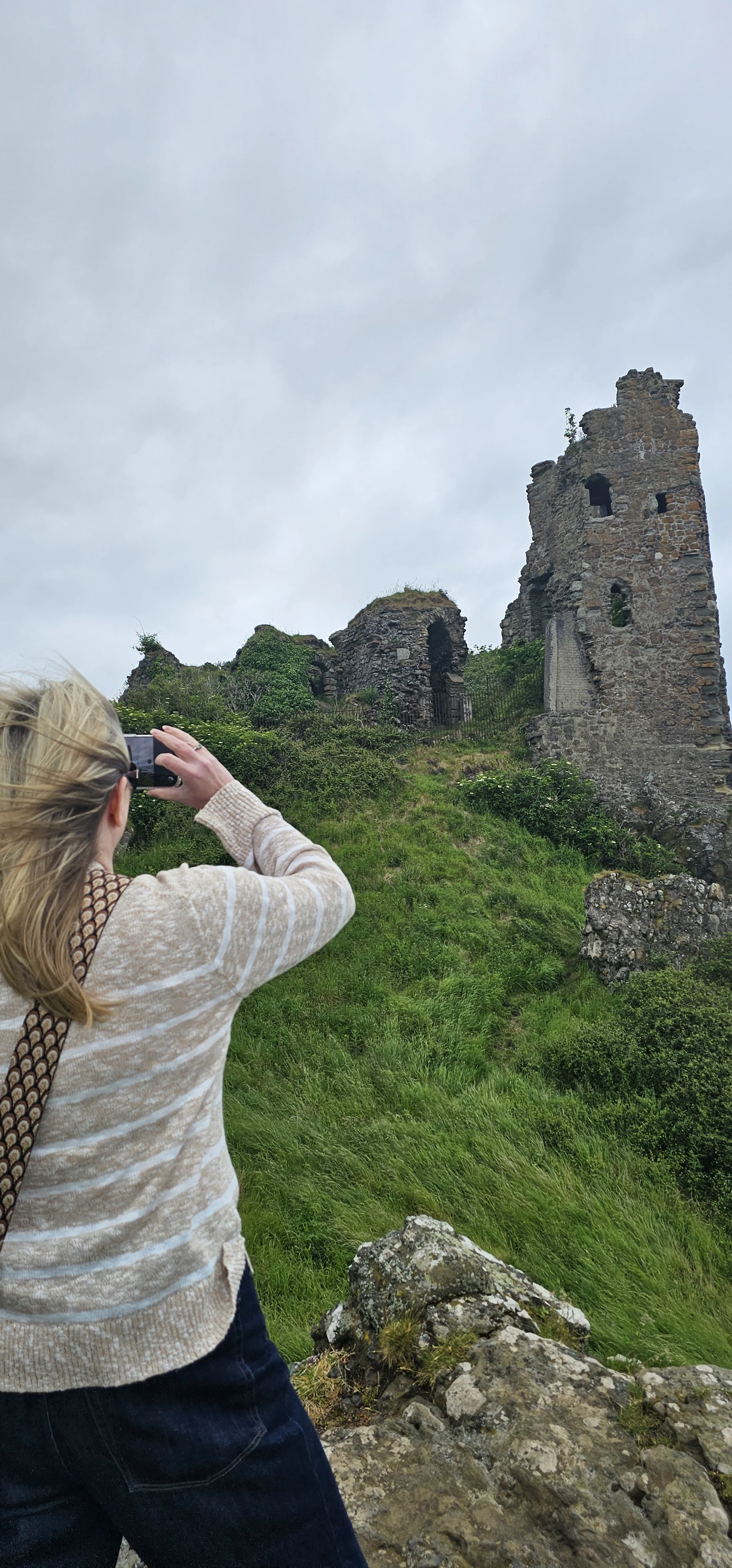 Visitor photographing ruined Scottish castle tower with smartphone on overcast day
