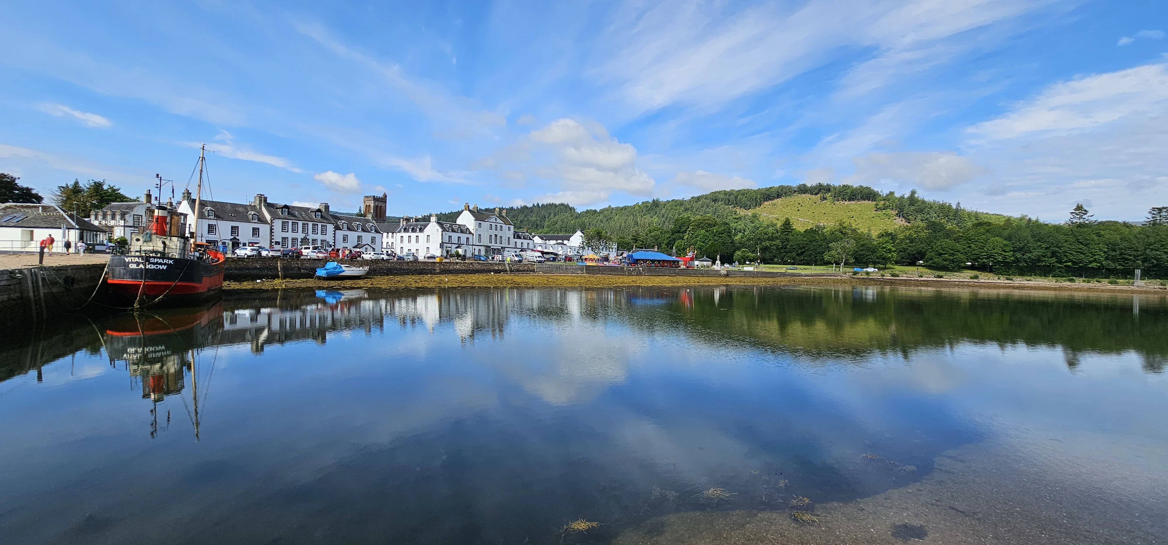 Scenic Scottish coastal village with white buildings, red boat, and forested hills reflected in calm water.
