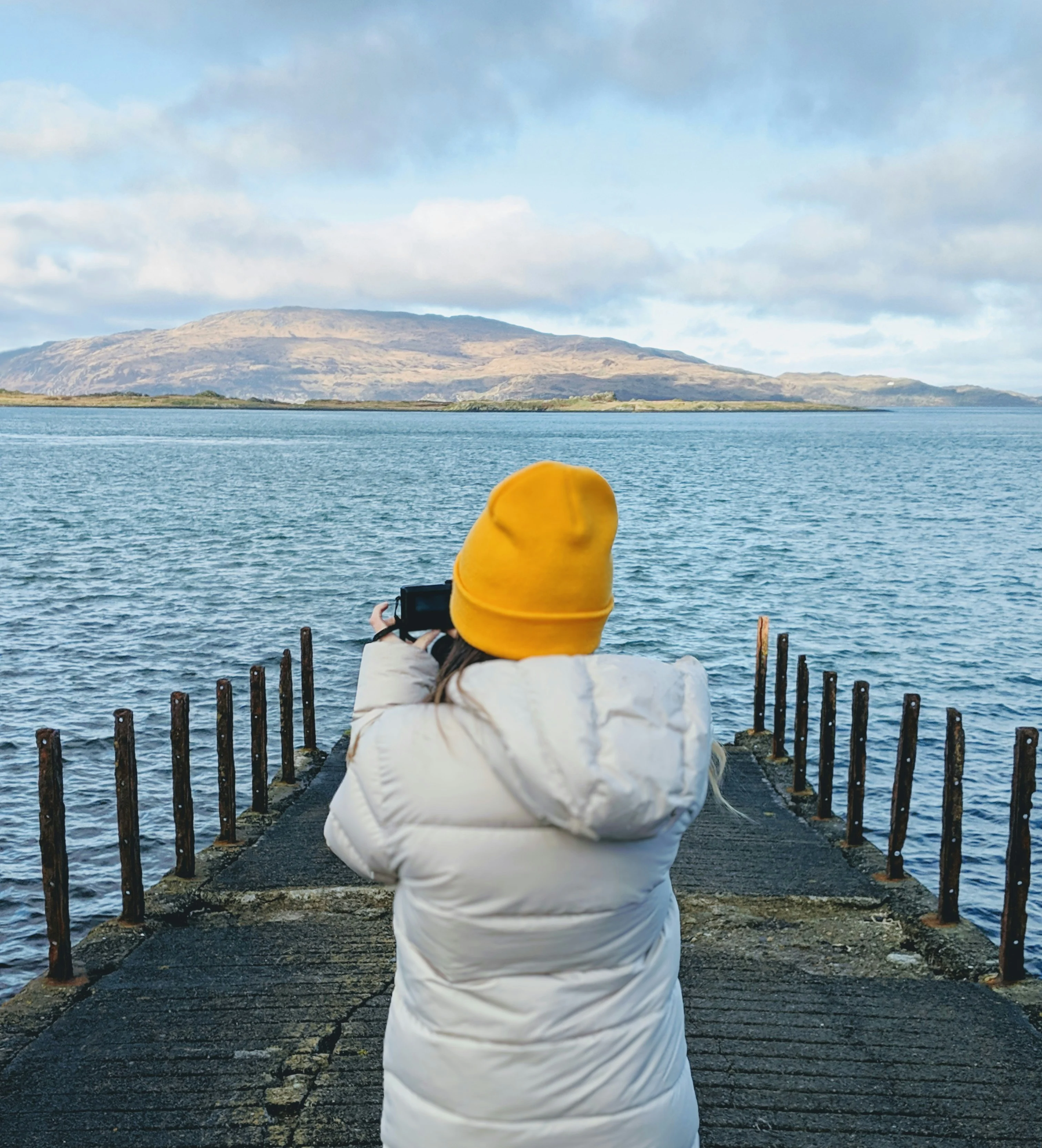Person in yellow beanie photographing Scottish mountain landscape across blue water