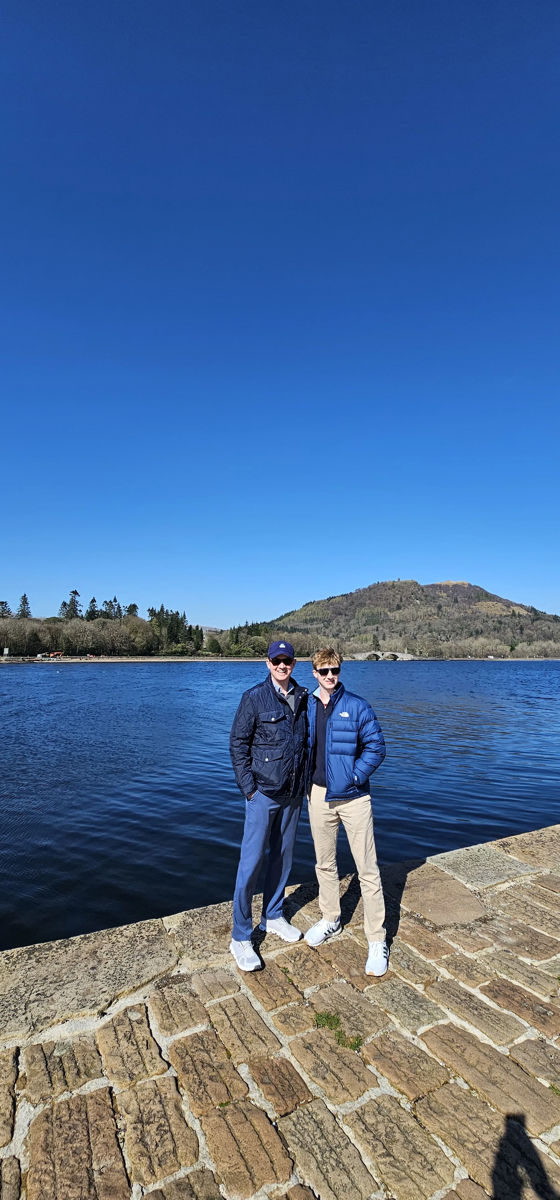 Two visitors posing by a Scottish loch with forested mountains in background.