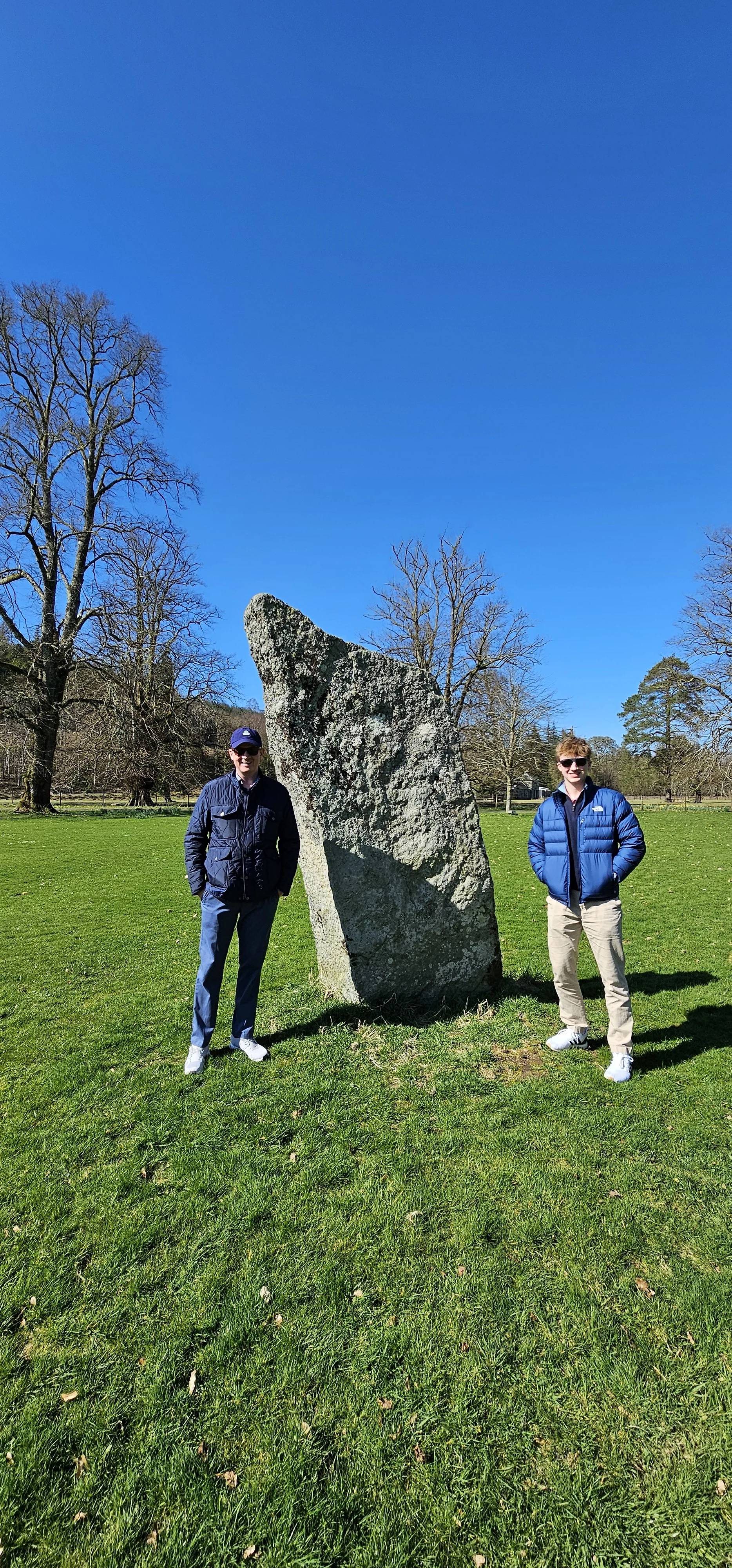 Two tourists stand beside an ancient Scottish standing stone in a green field.