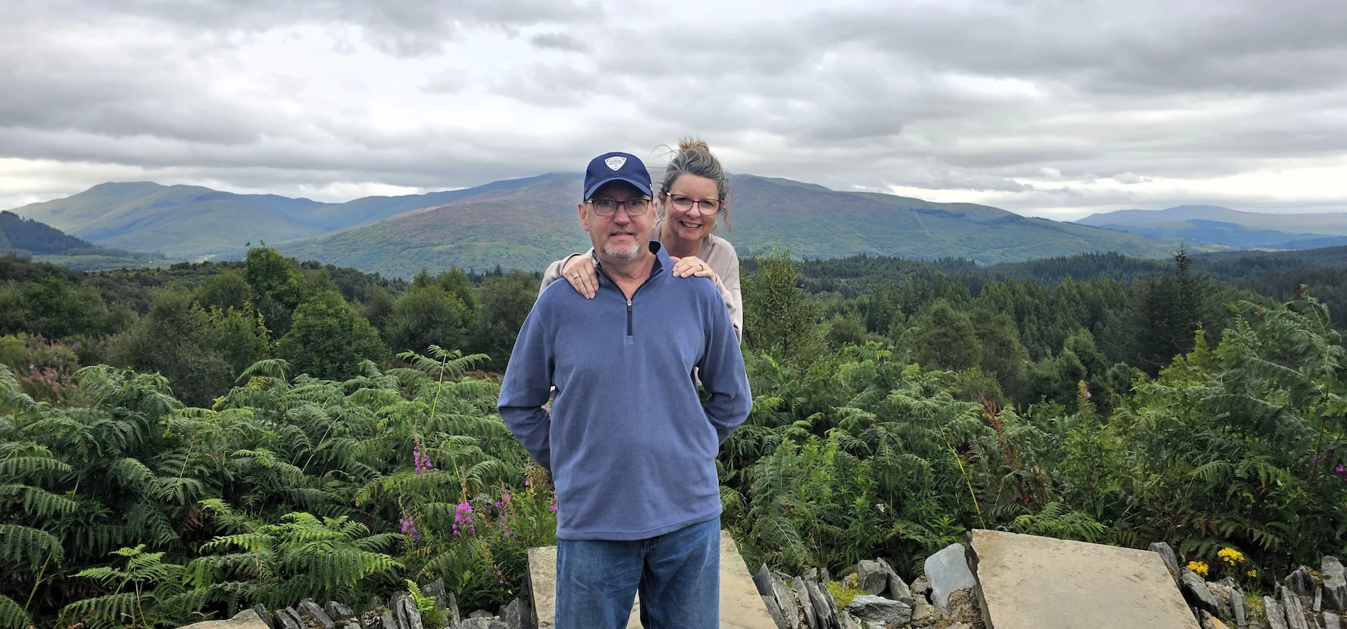 Couple posing at Scottish Highland viewpoint with mountains and forests in background.