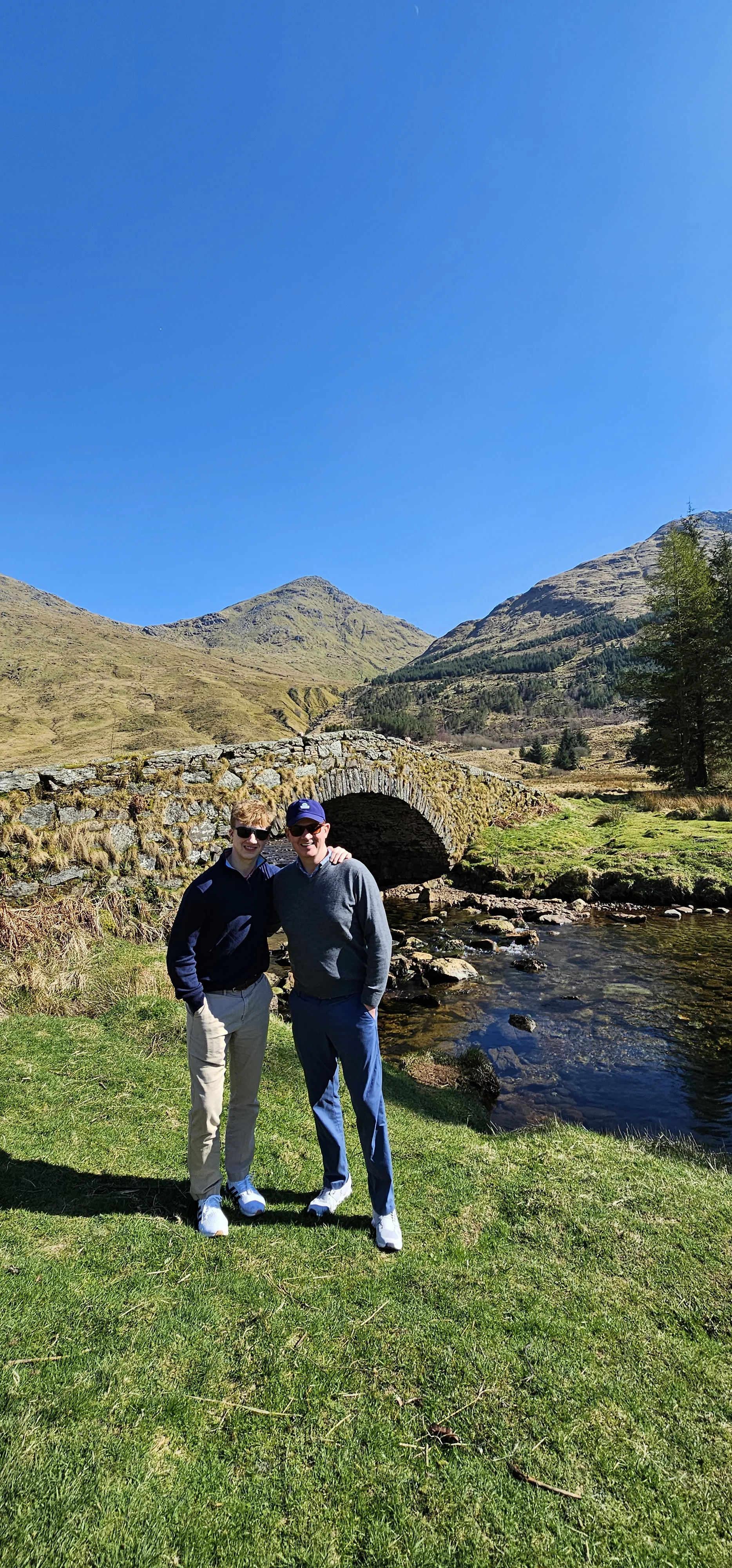 Two visitors posing by a historic stone bridge in a Scottish Highland valley