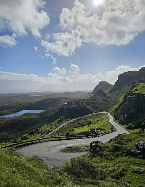 Winding mountain road through Scottish Highlands with loch and dramatic cliffs.