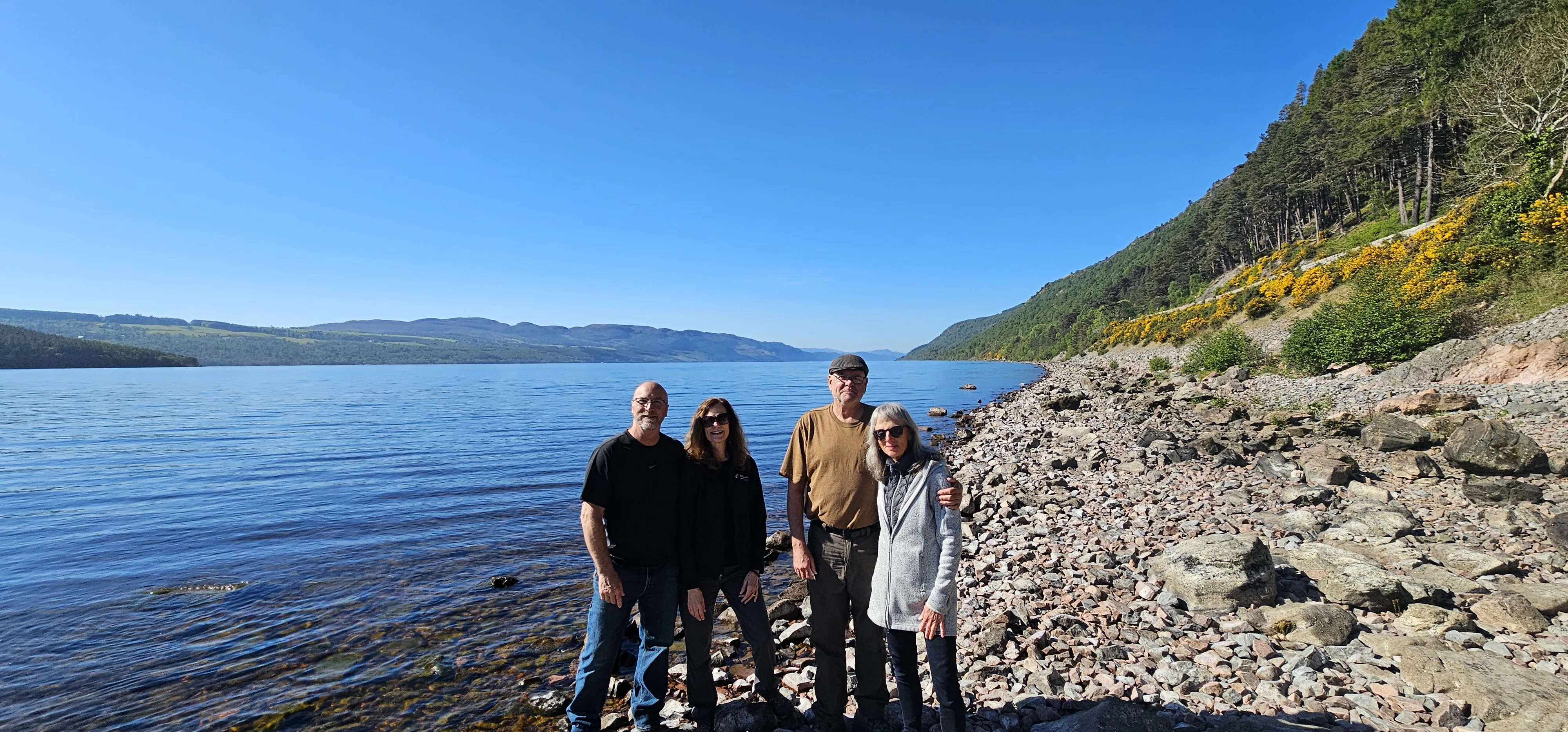 Four tourists posing on rocky beach beside Scottish loch under blue sky