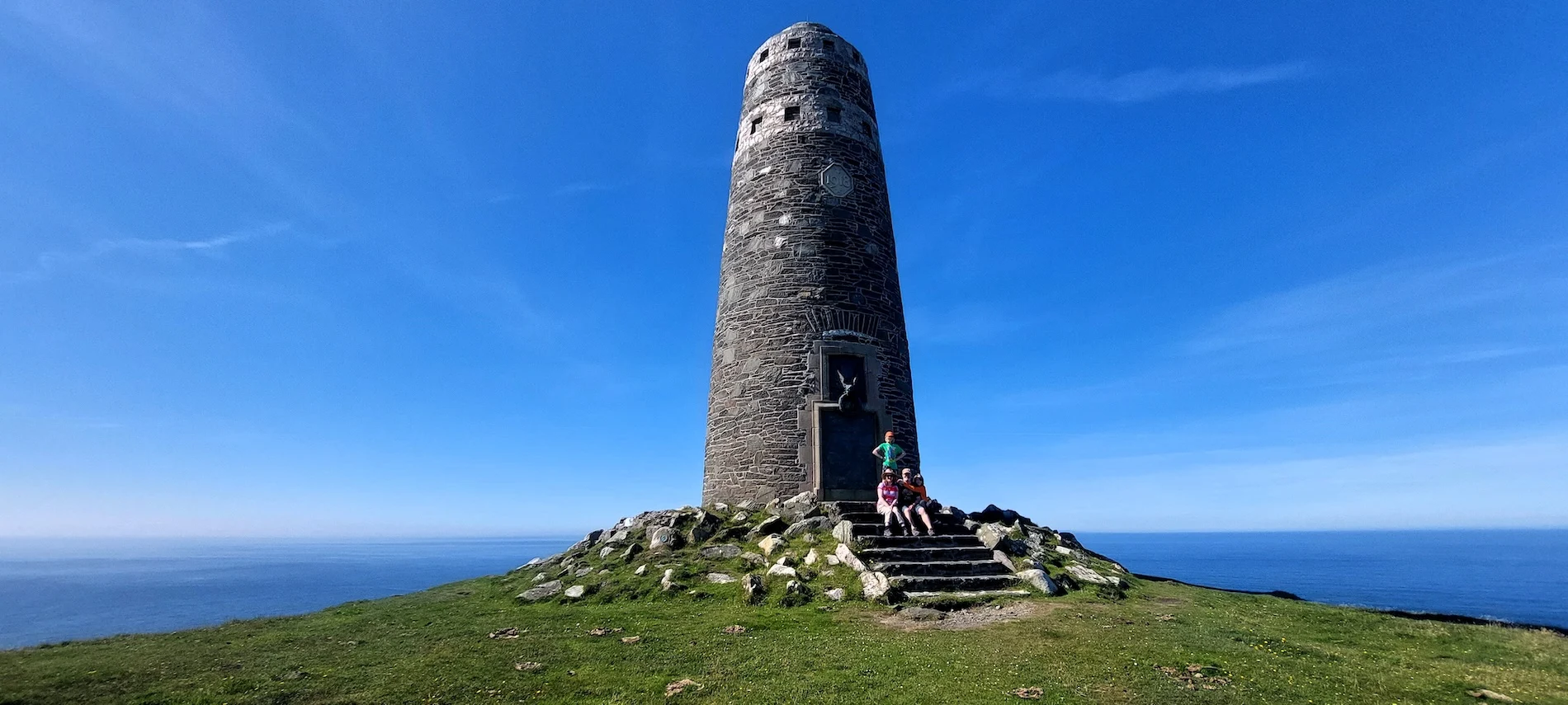 Stone tower on coastal hilltop with visitors and blue ocean backdrop