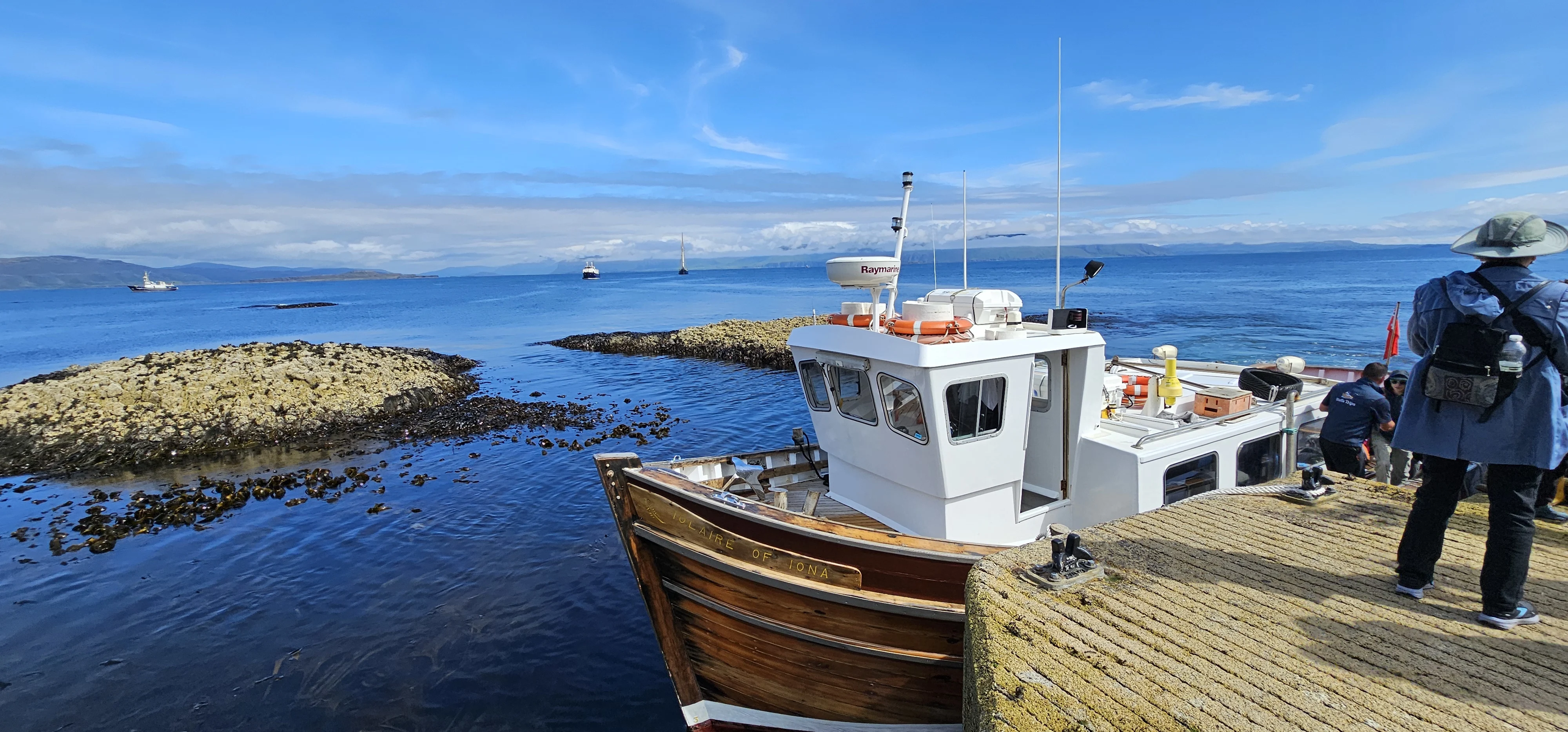 Fishing boats moored at a Scottish coastal pier with tourists and islands beyond