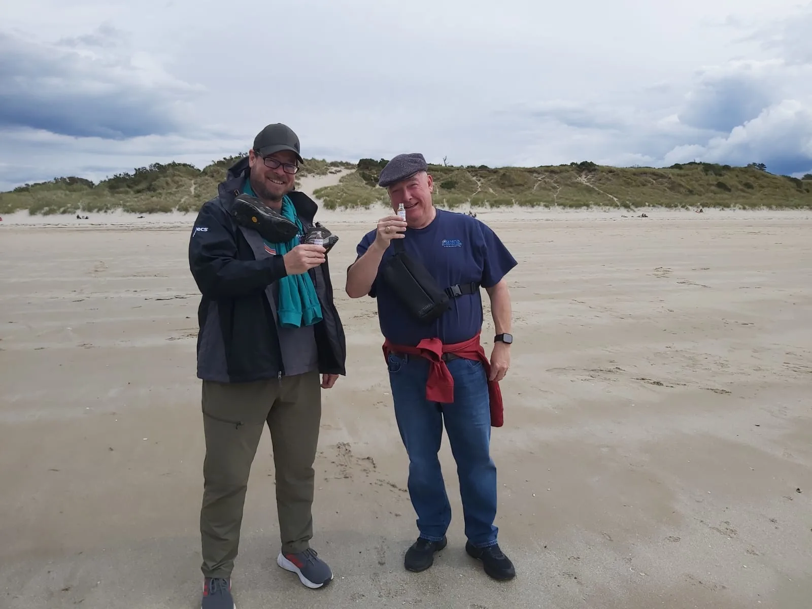 Two men smiling on a Scottish beach with dunes behind them.