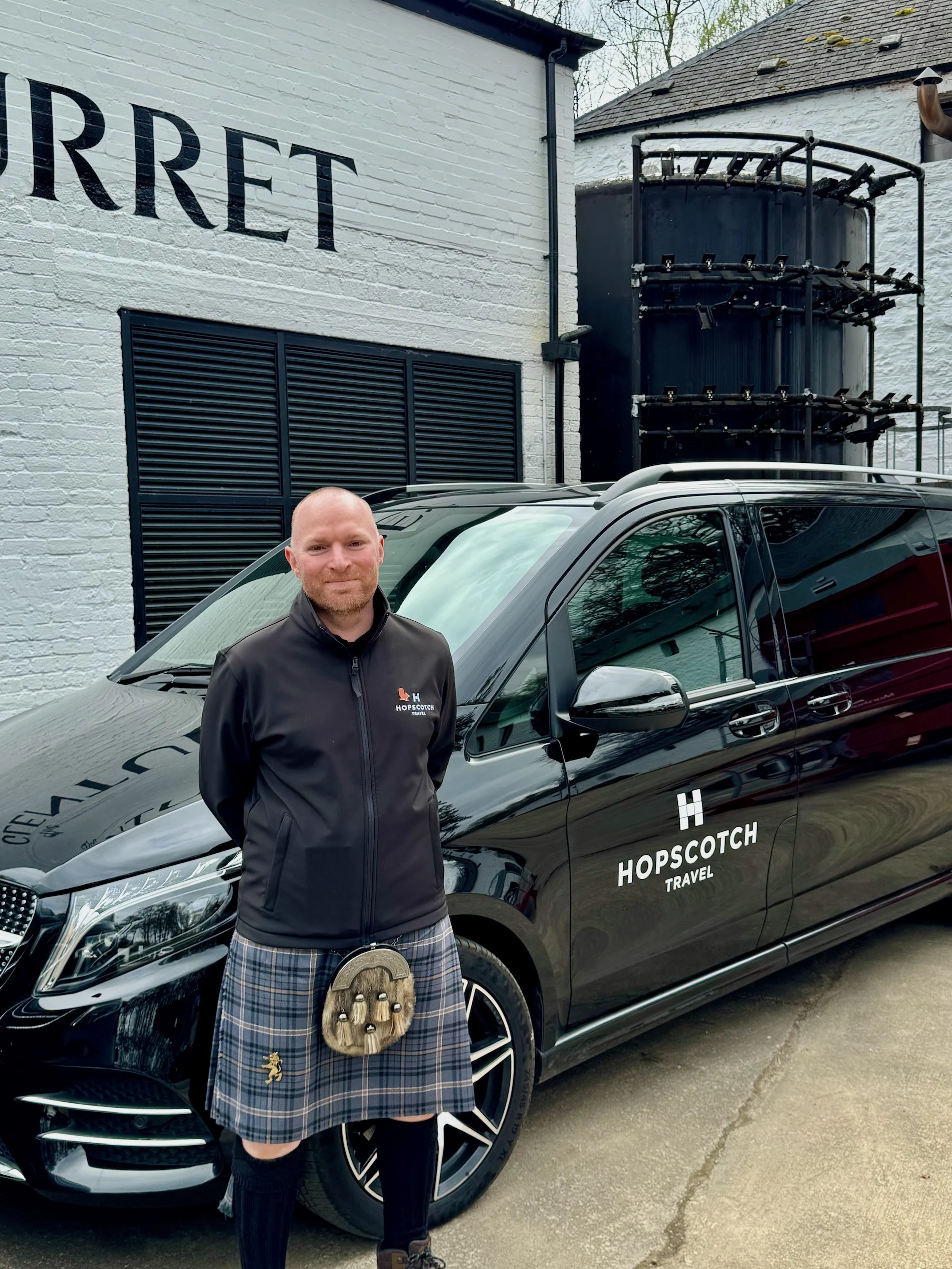 Tour guide in kilt standing beside Hopscotch Travel vehicle at Scottish distillery